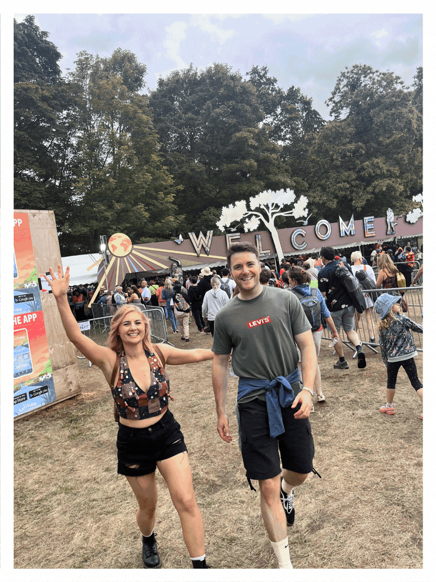 Two young people, a woman and a man, are smiling and holding hands while walking at an outdoor event. The woman has blonde hair, is wearing a black shorts and a patterned sleeveless top, and the man is wearing a green Levi's t-shirt, black shorts, and has a blue sweatshirt tied around his waist. They are in front of a large crowd near a welcome sign with trees in the background.