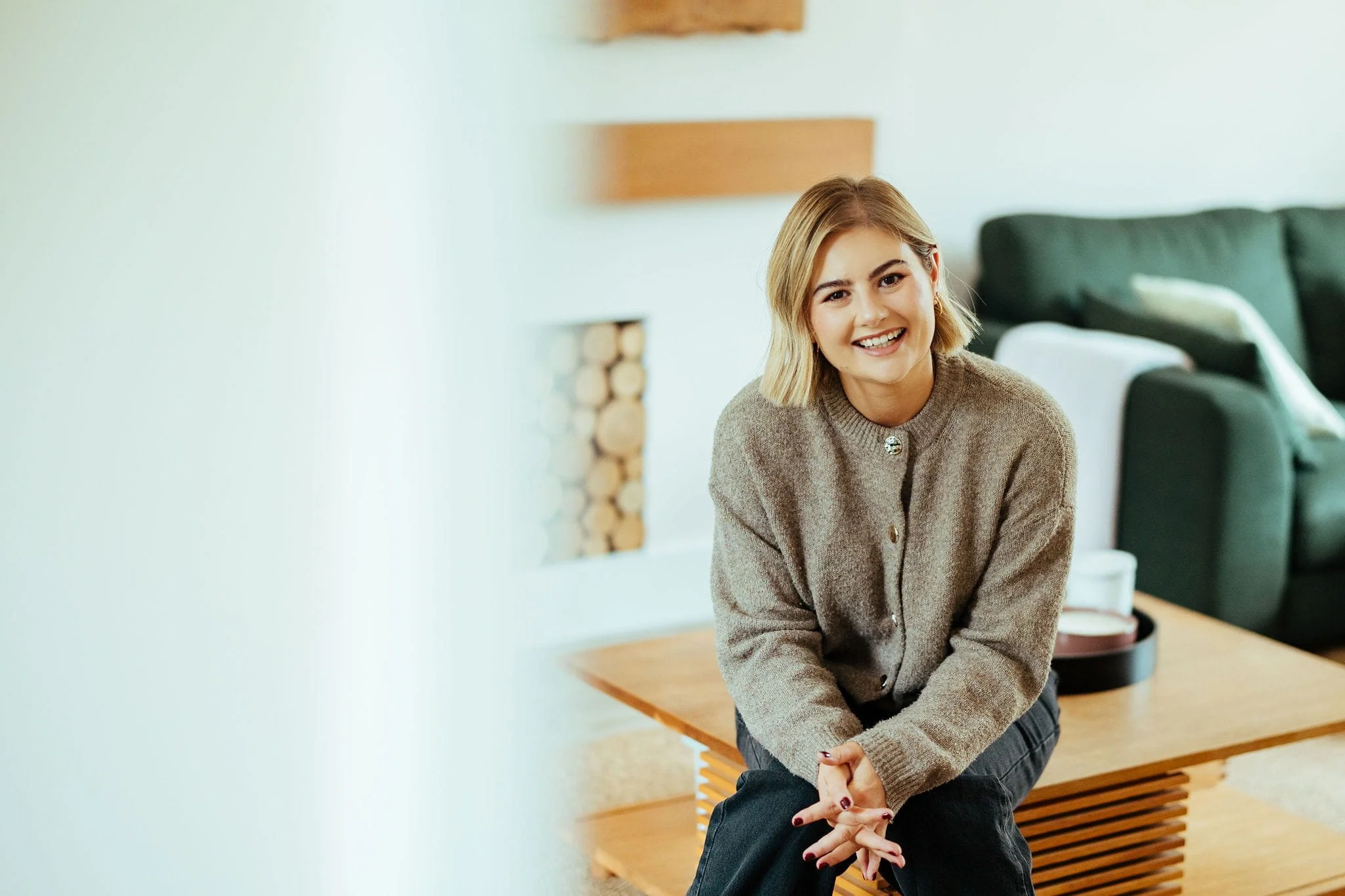 A young woman smiling, sitting on a wooden coffee table in a cozy living room with a green couch and cushions in the background.