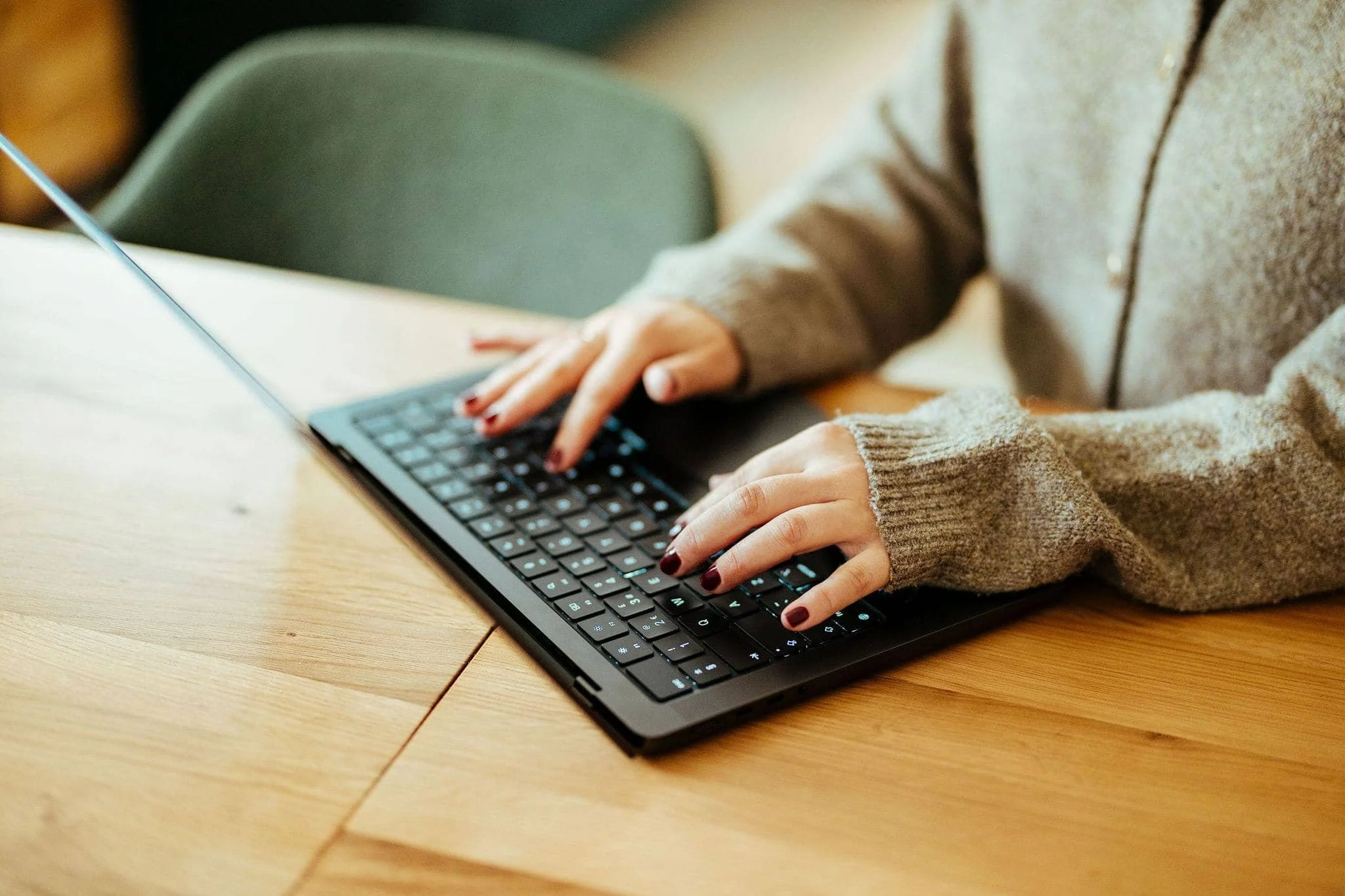 Person using a laptop keyboard, wearing a gray sweater, sitting at a wooden table with a green chair nearby.