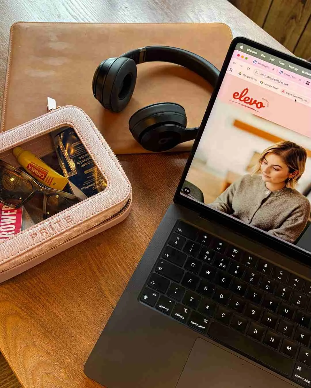 flatlay of a macbook, with Elevo website in view, headphones and a bag of personal items such as sunglasses and handcream