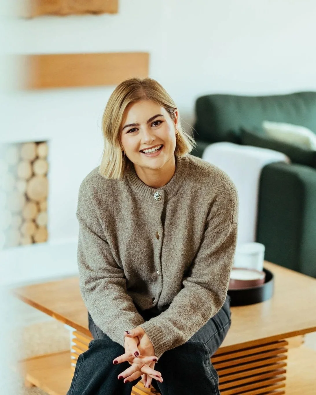 Squarespace website designer Elie Hicks smiling, sat on a table with knees and hands together. Wearing a cardigan and jeans.
