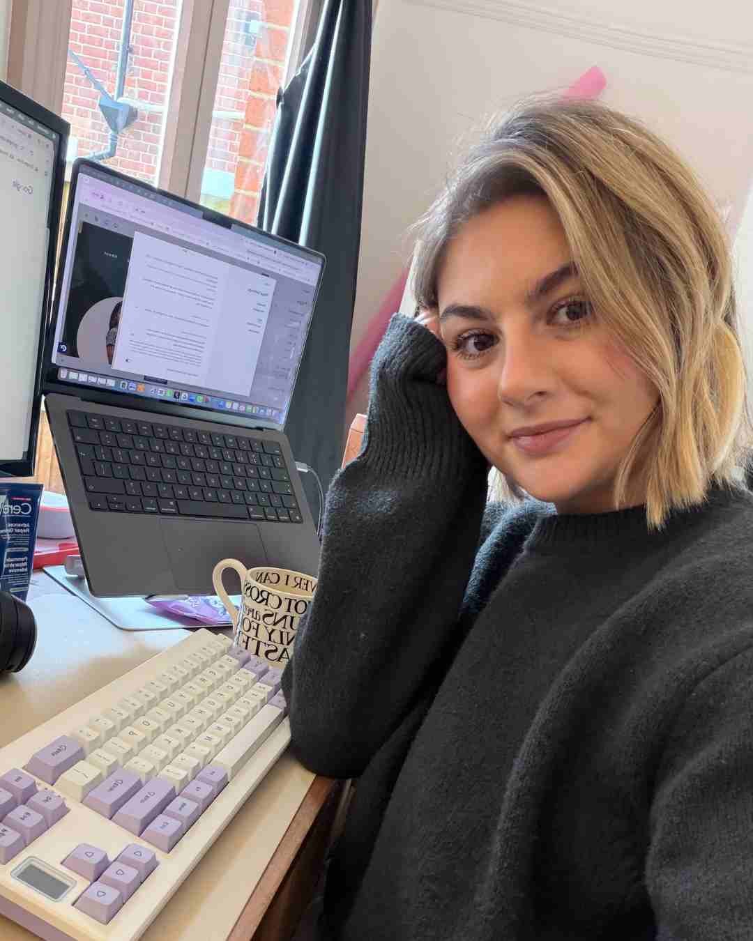 woman sat at her desk with keyboard, laptop and cup of tea, wearing a grey jumper smiling at the camera