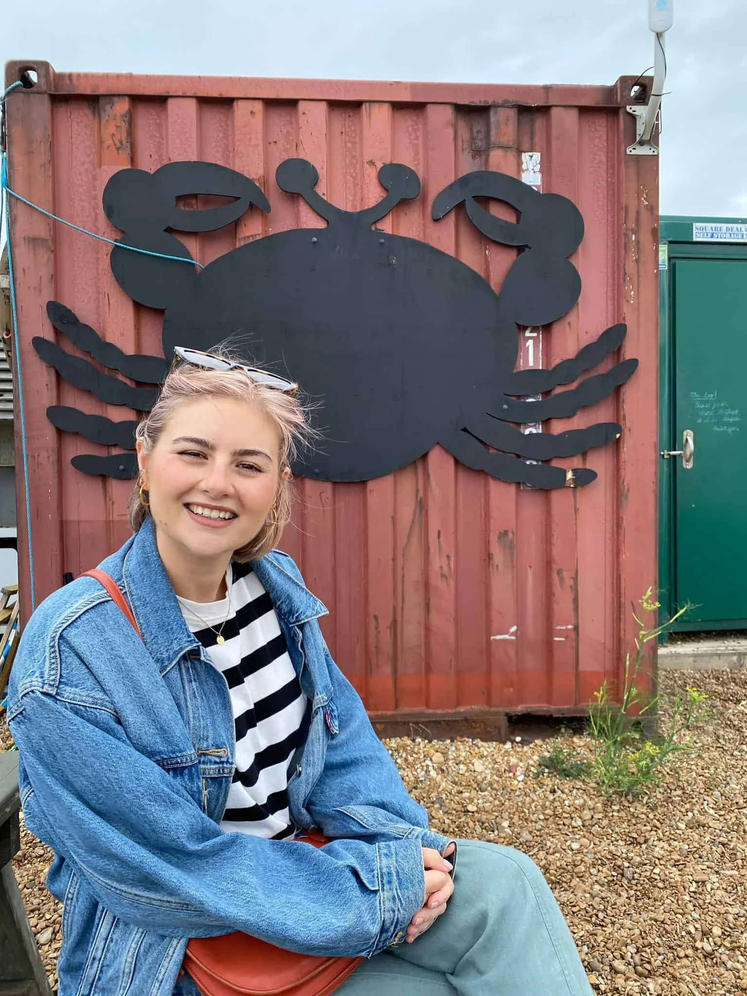 A smiling young woman with blonde hair, wearing a denim jacket and black-and-white striped shirt, sitting on a bench in front of a large black crab silhouette on a red metal container.