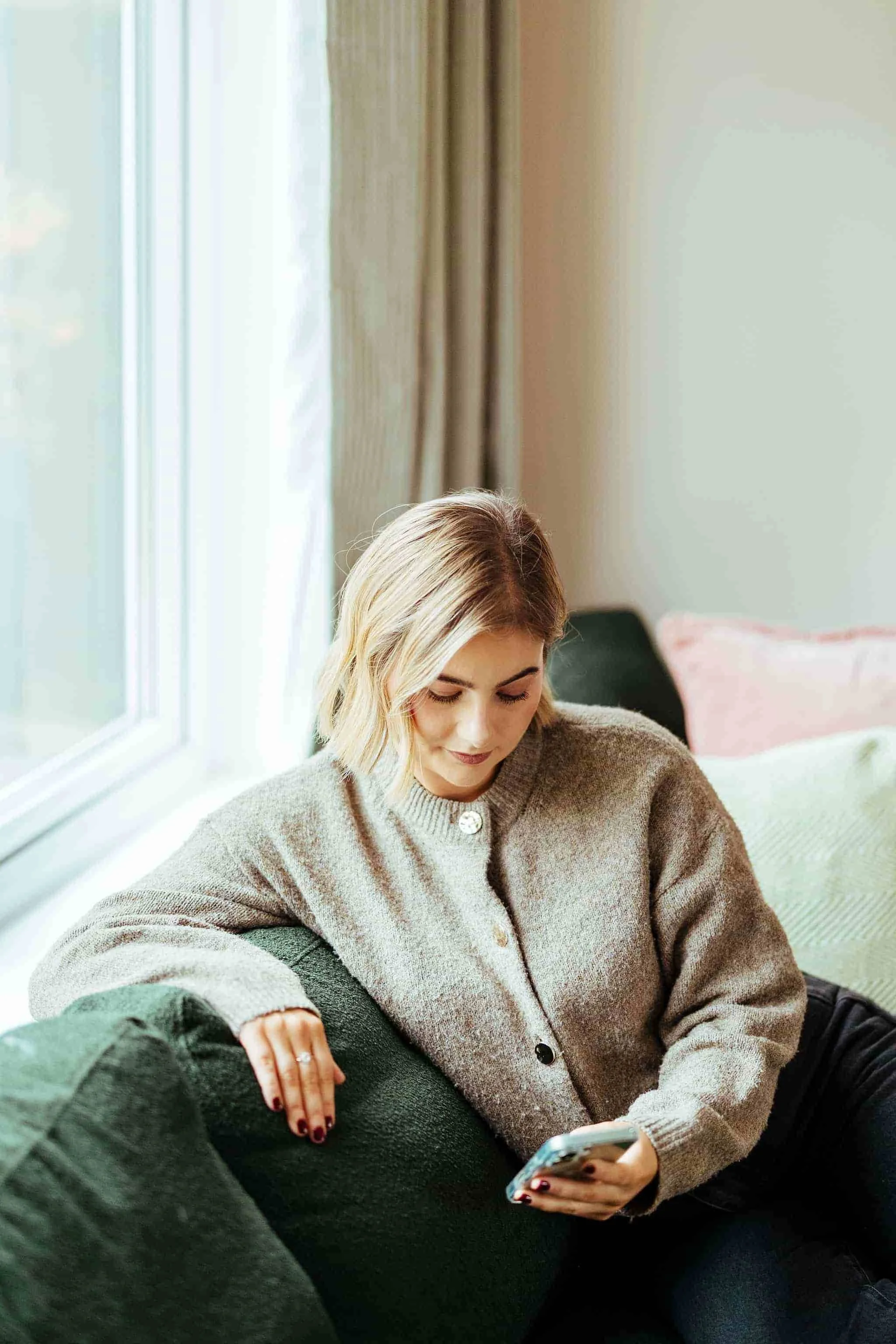 Squarespace Website Designer with short blonde hair sitting on a green couch near a window, looking at her phone and wearing a beige sweater.