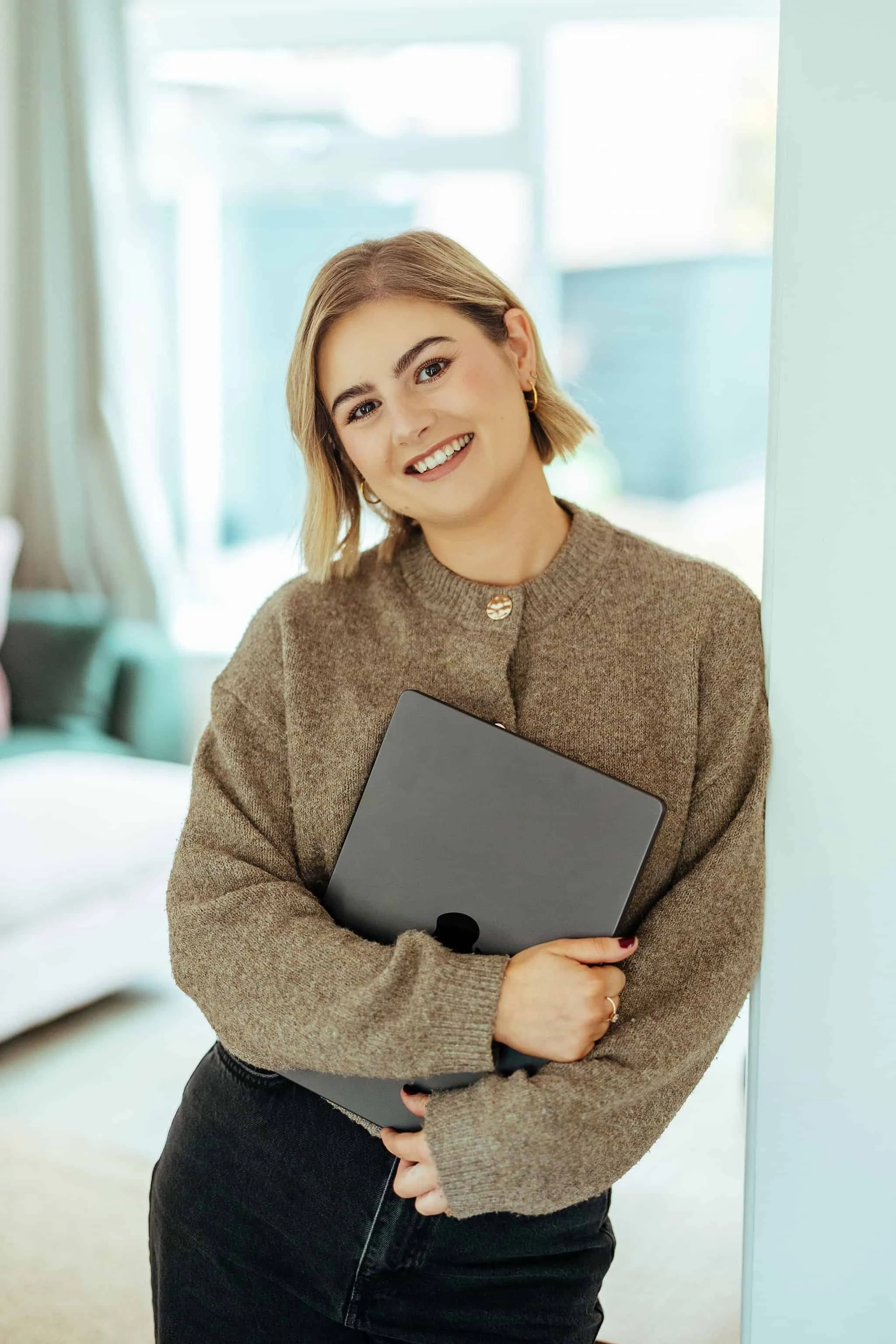 A young woman with short blonde hair holding a tablet, smiling, wearing a brown sweater and earrings, standing in a well-lit room.