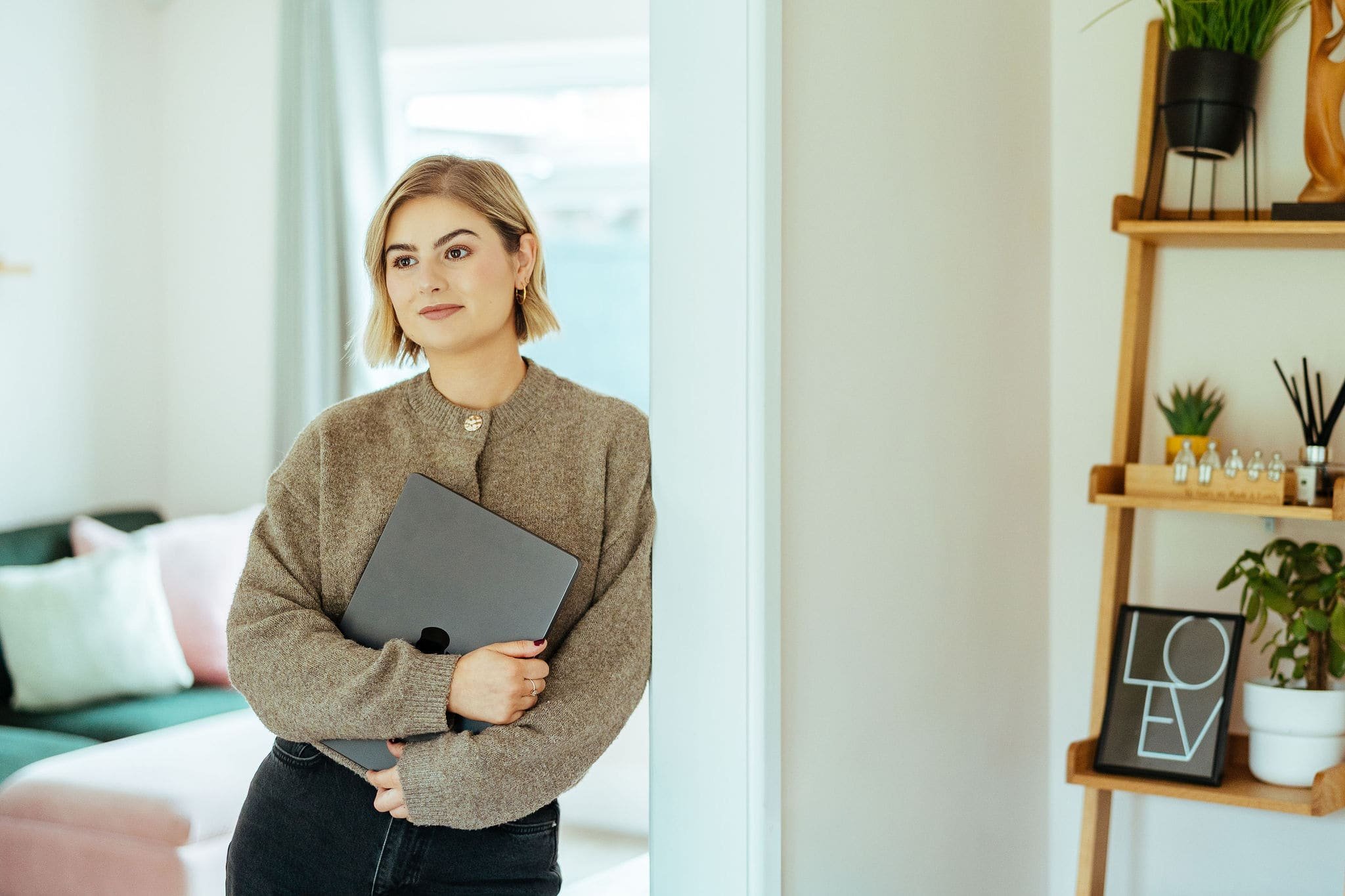 A young woman with shoulder-length blonde hair, wearing a brown sweater, holding a closed laptop, standing in a cozy, well-lit room with a green sofa and decorative items on a wooden shelf.