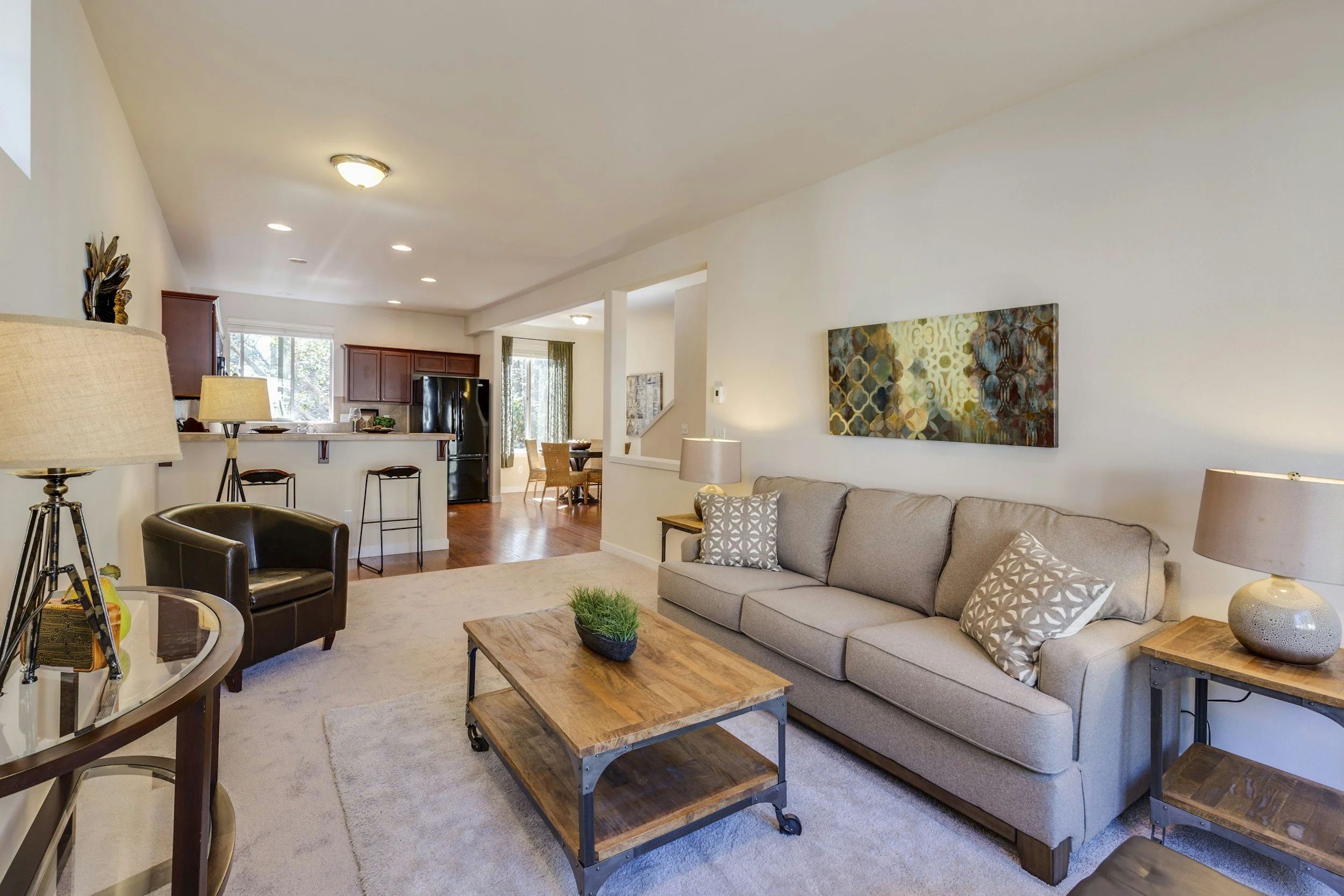 Living room with beige sofa, wooden coffee table, black armchair, and various lamps, opening to kitchen and dining area with black refrigerator and wooden dining table.