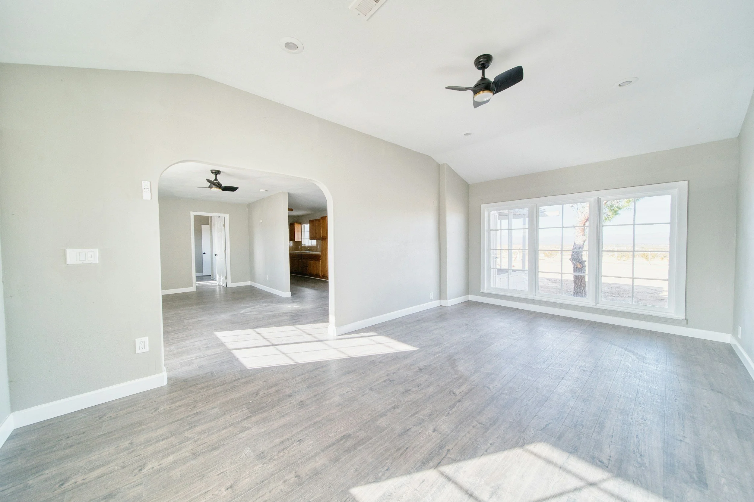 Bright living room with large windows, light gray walls, gray wood flooring, and ceiling fans, with an open view into the kitchen.