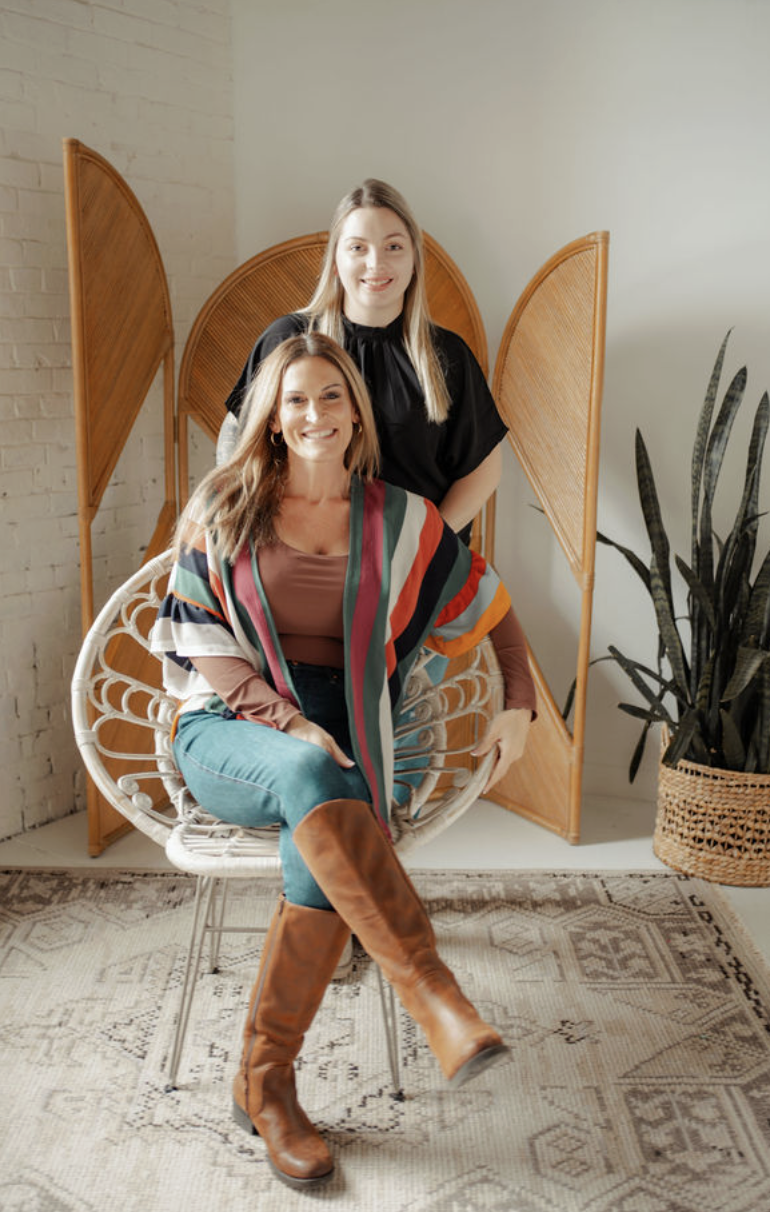 Two women posing indoors, with one seated on a wicker chair and the other standing behind her. The seated woman wears jeans, a colorful striped shawl, brown shirt, and tall brown boots. The standing woman wears a black top. There is a wooden room divider, a potted plant, and a patterned rug in the background.