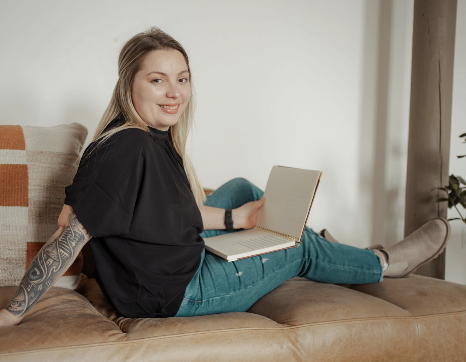 A young woman with blond hair and a tattoo on her arm is sitting on a tan leather couch, smiling, and holding an open notebook, with her legs stretched out and relaxed in a cozy living room.