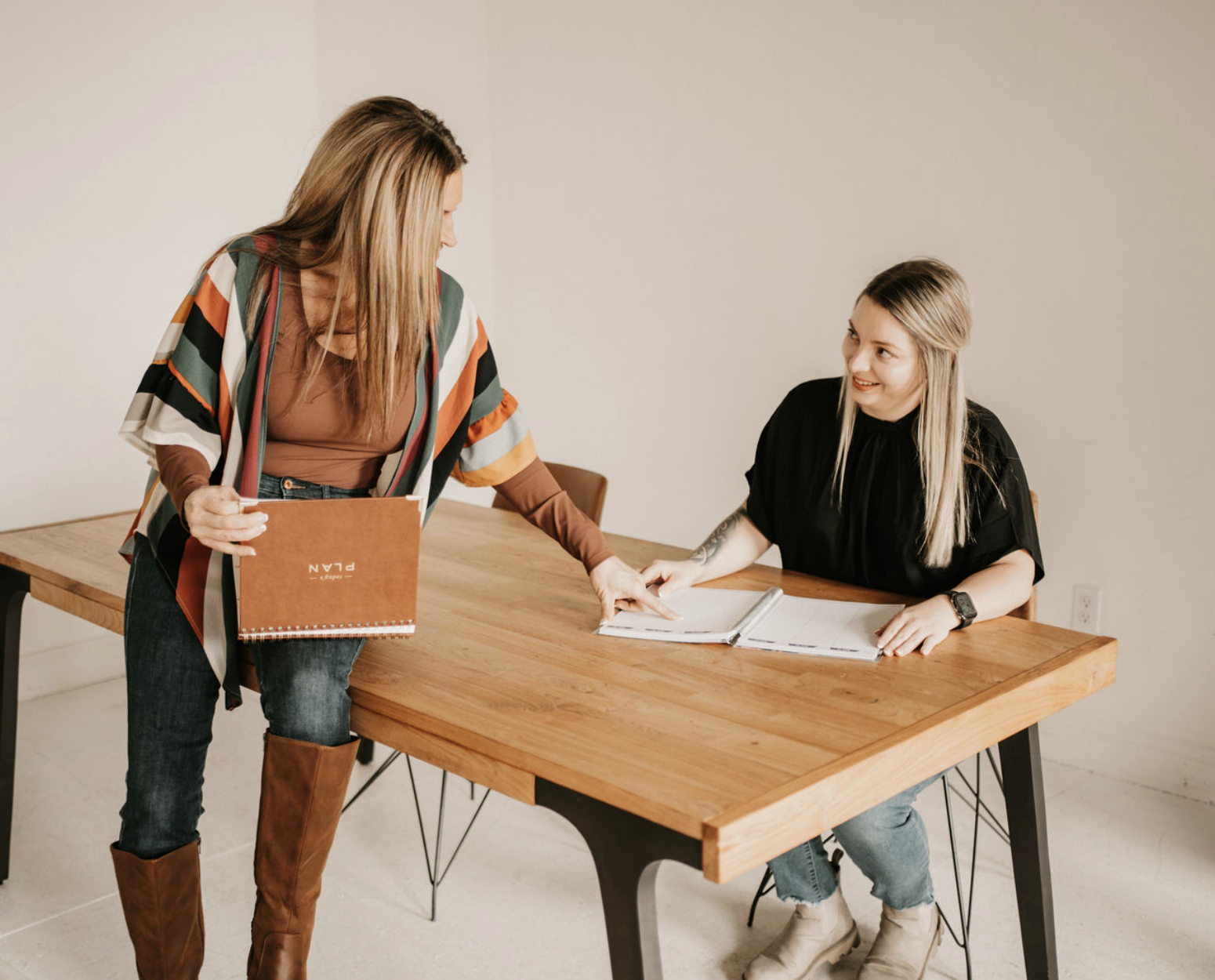 Two women in a meeting, one standing and pointing at a notebook, the other sitting at a wooden table, smiling, with open documents.