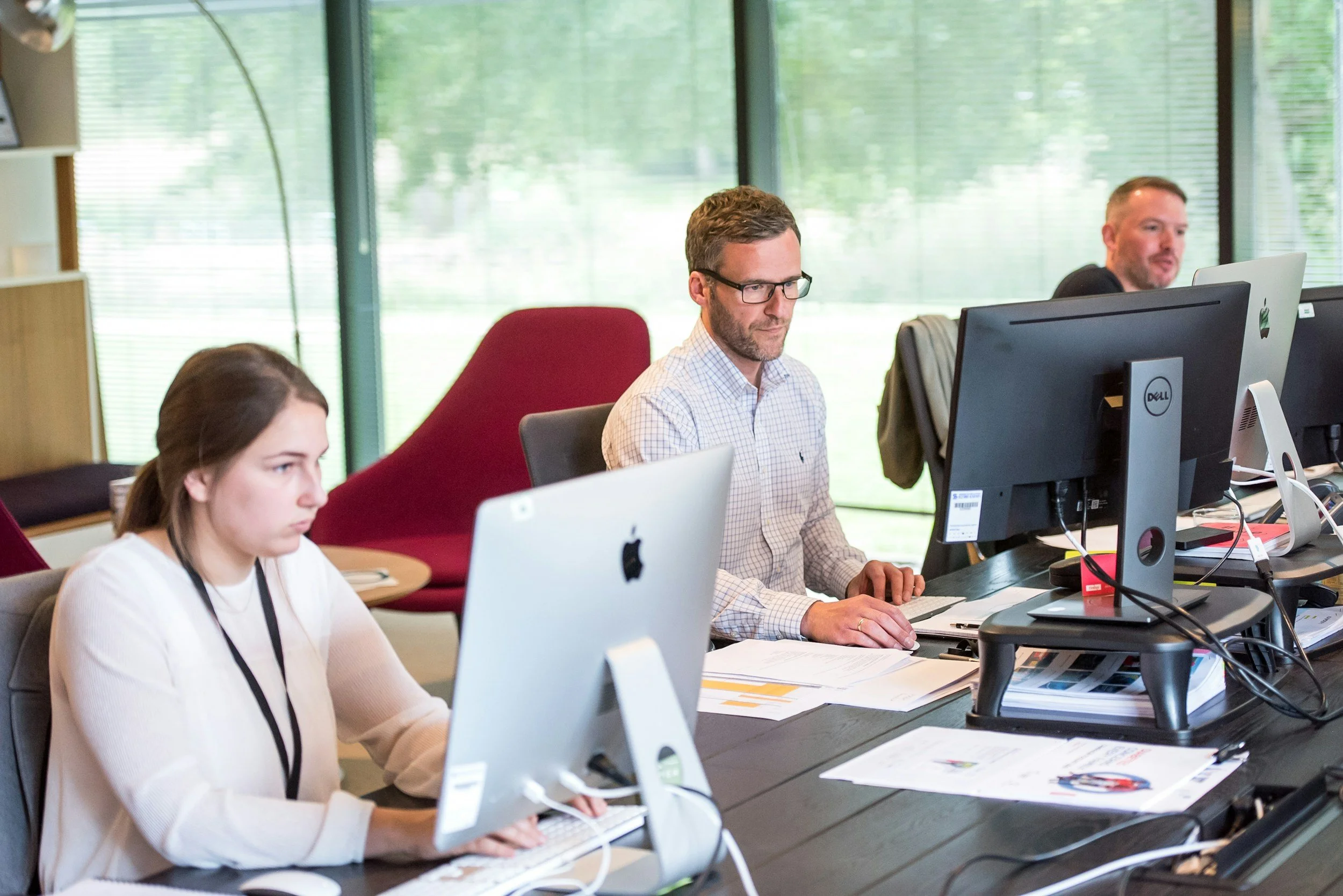 Three people working at their desks in an office with large windows and green trees outside. The woman on the left has brown hair and is working on an iMac. The man in the middle with glasses has a beard and is working on a desktop computer. The man on the right has short hair and is smiling while working on a desktop computer.