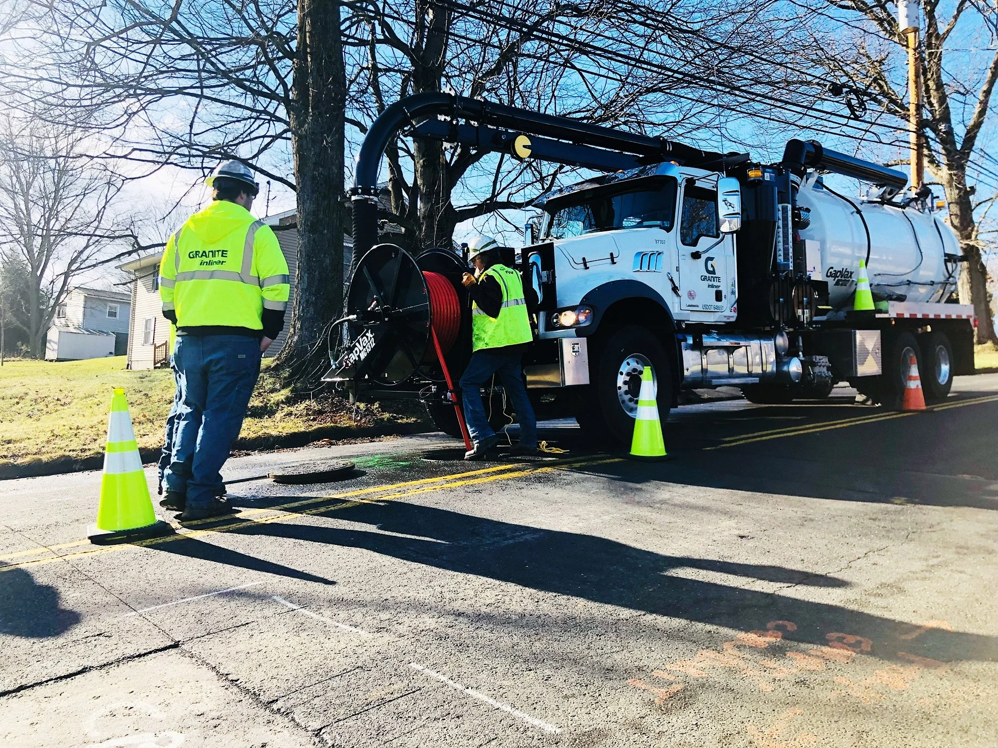 Three utility workers in bright yellow safety vests and helmets working around a large GapVax MC combo truck near Columbus, Tennessee. They are cleaning a sewer line by putting the hose into a manhole and running high-pressure water through the line.