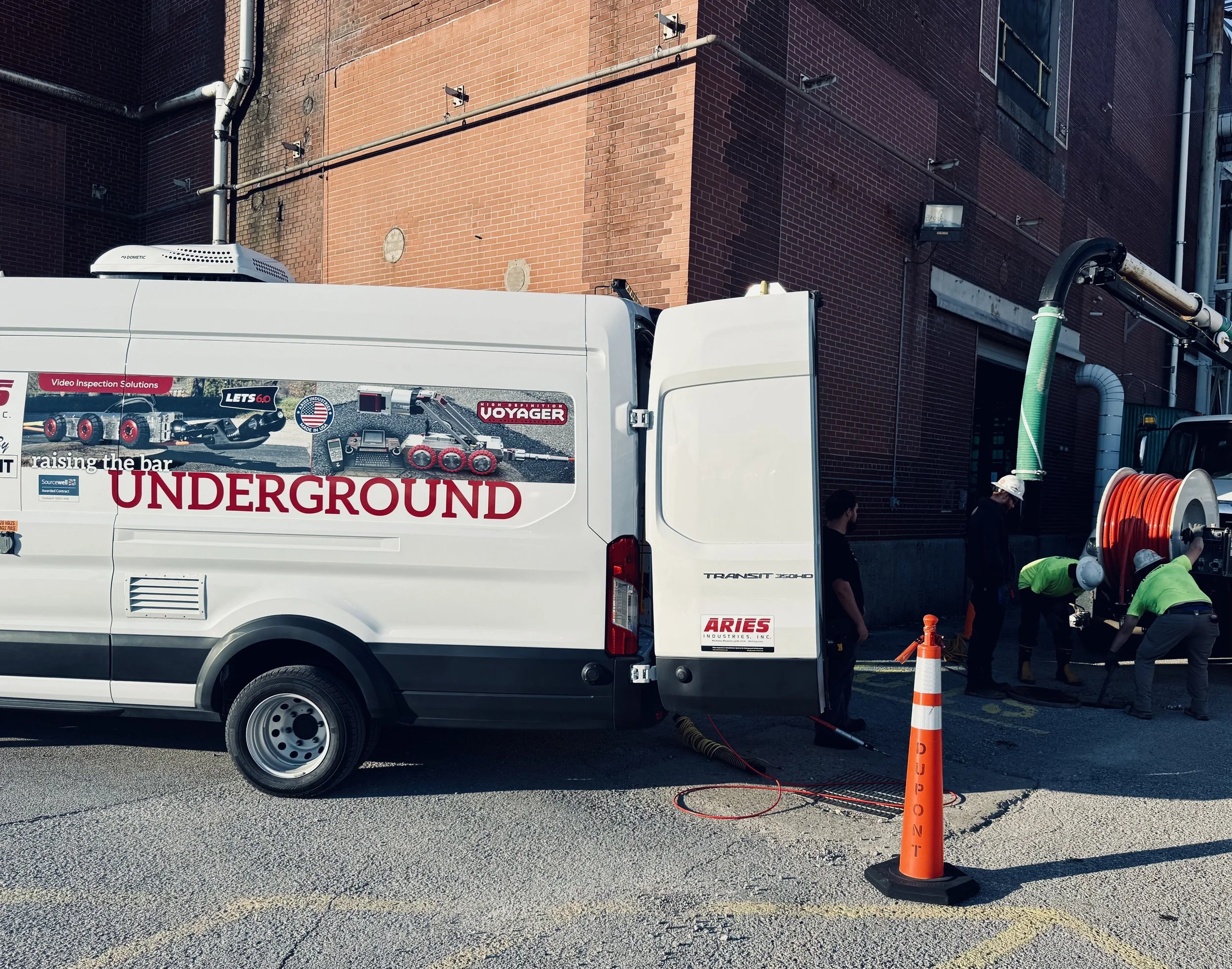 Utility workers in green and black uniforms working on underground inspecting a mainline sewer pipe with an Aries Voyager Mainline System in a Ford Transit Van parked near a brick building, with orange traffic cone and equipment around.