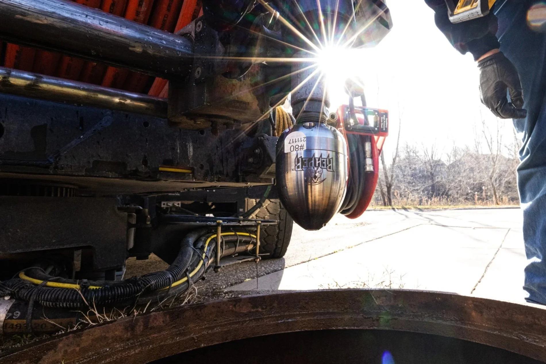 Close-up of a vacuum truck with reaper nozzles hanging from it and hoses visible, sunlight shining through the background, and a person wearing jeans and gloves standing nearby.
