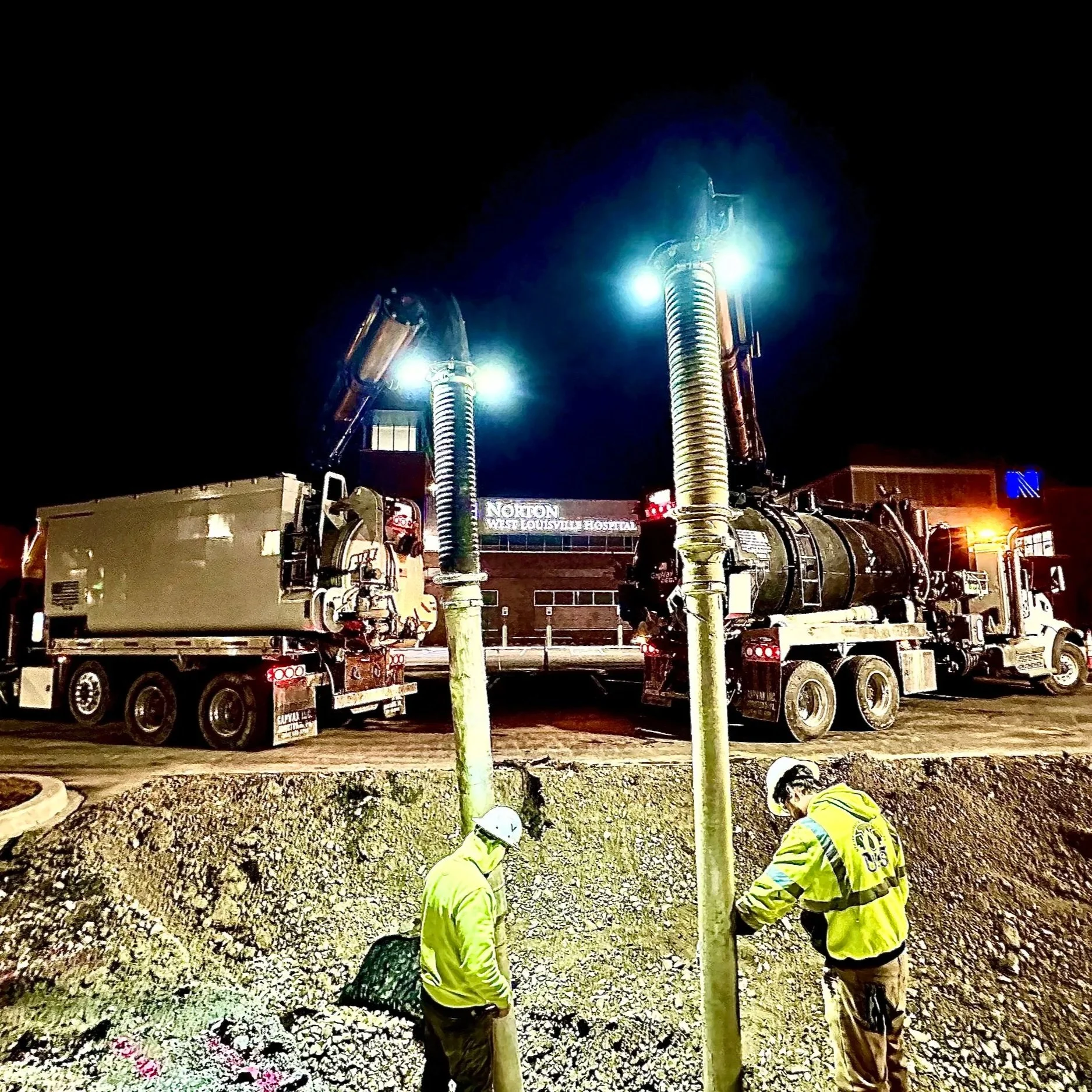 Two construction workers wearing safety gear working on underground pipes at night, with large vacuum trucks and bright lights illuminating the scene outside Norton hospital building in Louisville, KY
