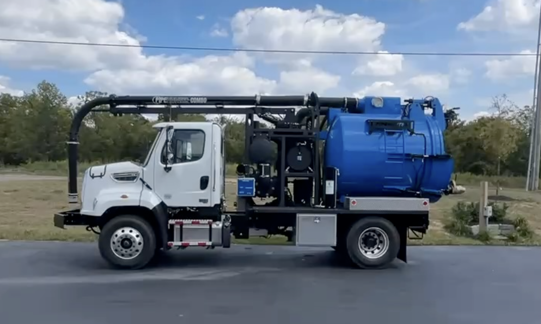 A white and blue PipeHunter mini hydro-excavator vacuum truck parked on a road with grassy area and trees in the background under a partly cloudy sky near Brazil, Indiana.