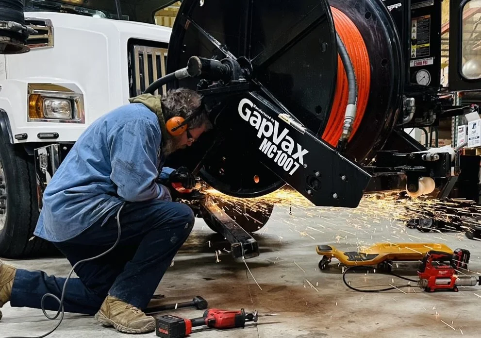 an expert mobile service techniciation wearing safety gear, including ear protection and gloves, kneeling and welding the front hose reel of a GapVax MC vac truck at a service call in Indianapolis, Indiana.