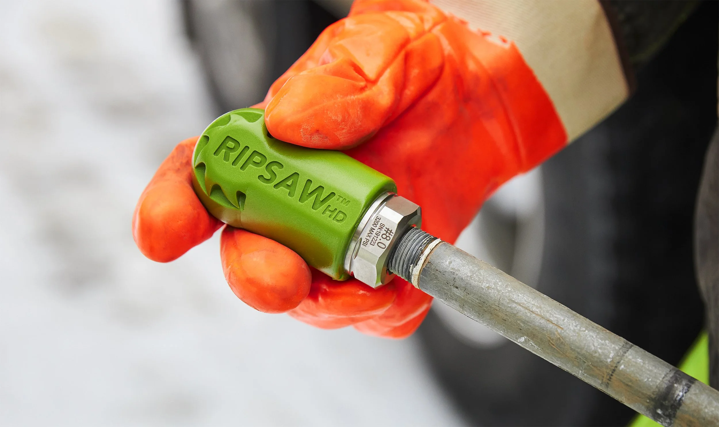 Close-up of a person's hand wearing an orange work glove, holding a green ripsaw hydro excavation nozzle attached to a lance. 