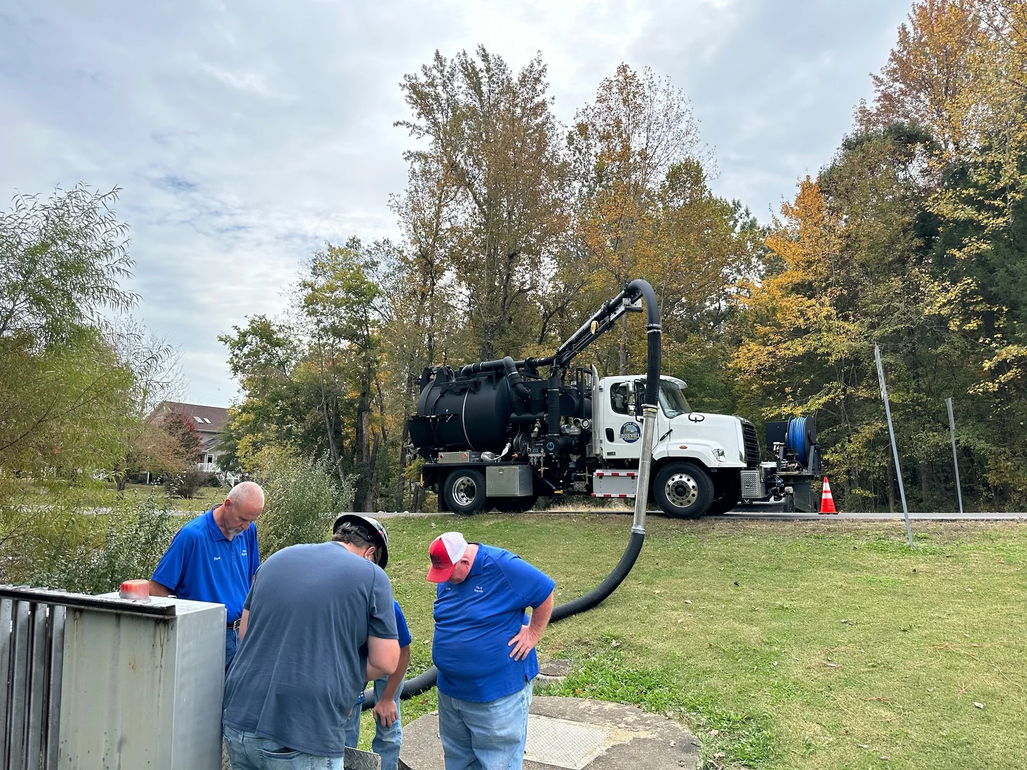 Three workers in blue shirts cleaning a lift station with a  vacuum truck parked on a grassy area near trees with autumn foliage.