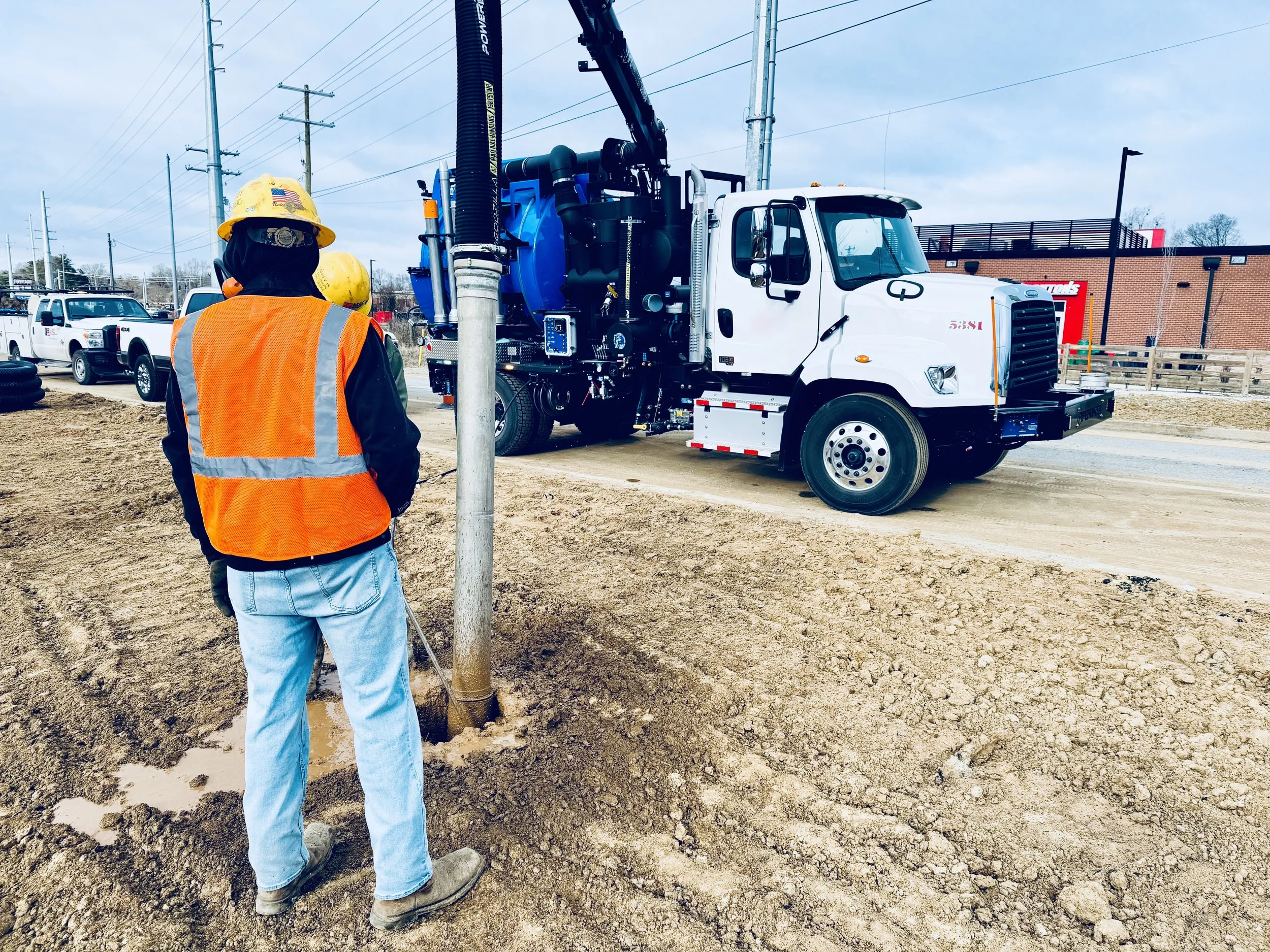 A construction worker wearing an orange safety vest and yellow helmet operates a PipeHunter vacuum truck at a construction site. 