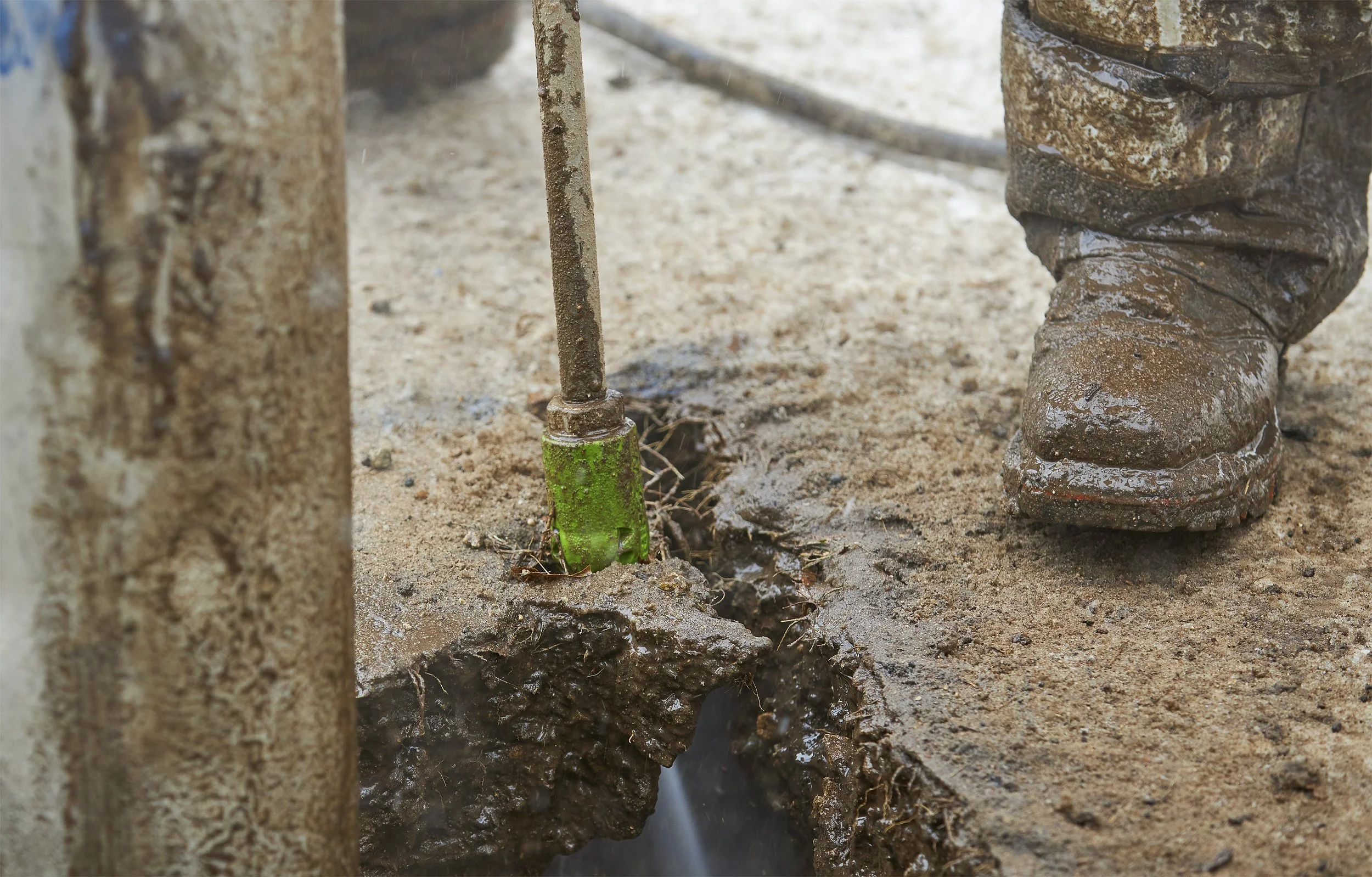 Close-up of a worker's muddy boot next to a hole he is digging using hydro excavation. he is using a green ripsaw nozzle to dig with high pressure water and safely uncover utilities.
