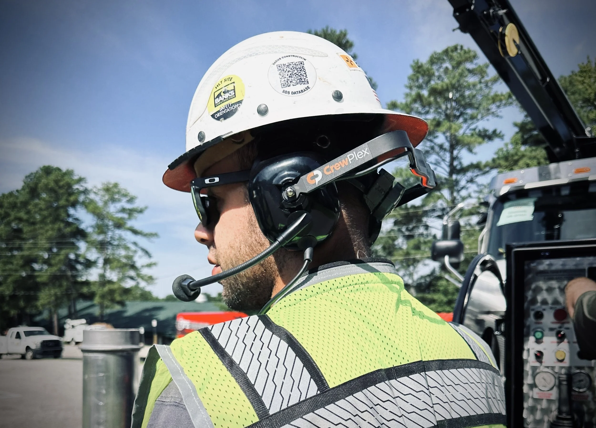 Construction worker wearing a white hard hat, black sunglasses, a Crewplex wireless headset sold by 502 Equipment with microphone, and a high-visibility safety vest, working outdoors with equipment and trees in the background in Louisville, KY