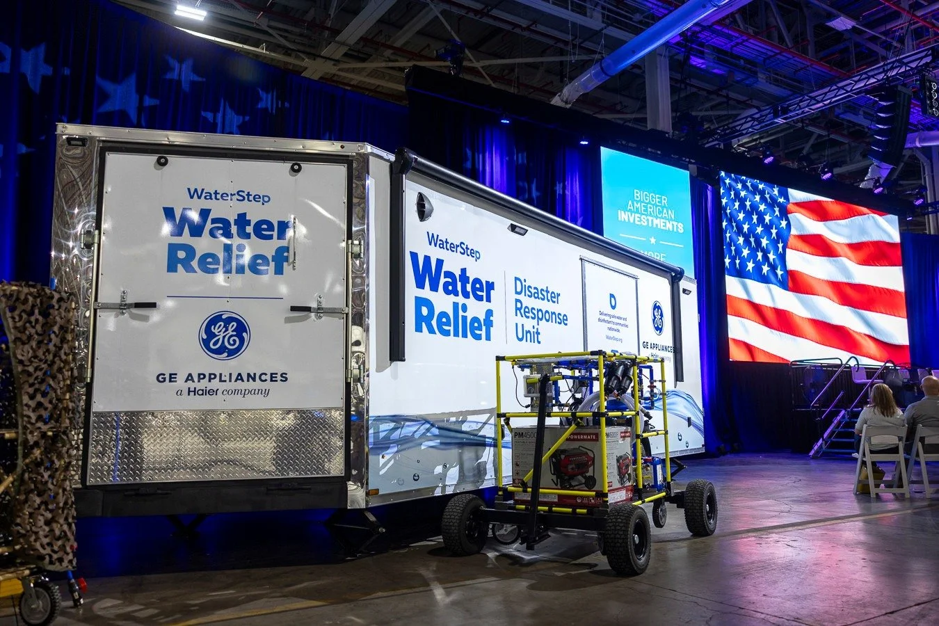A mobile water relief trailer unit by Waterstep in partnership with GE Appliances at an indoor event, with a large American flag screen and a blue presentation screen in the background.