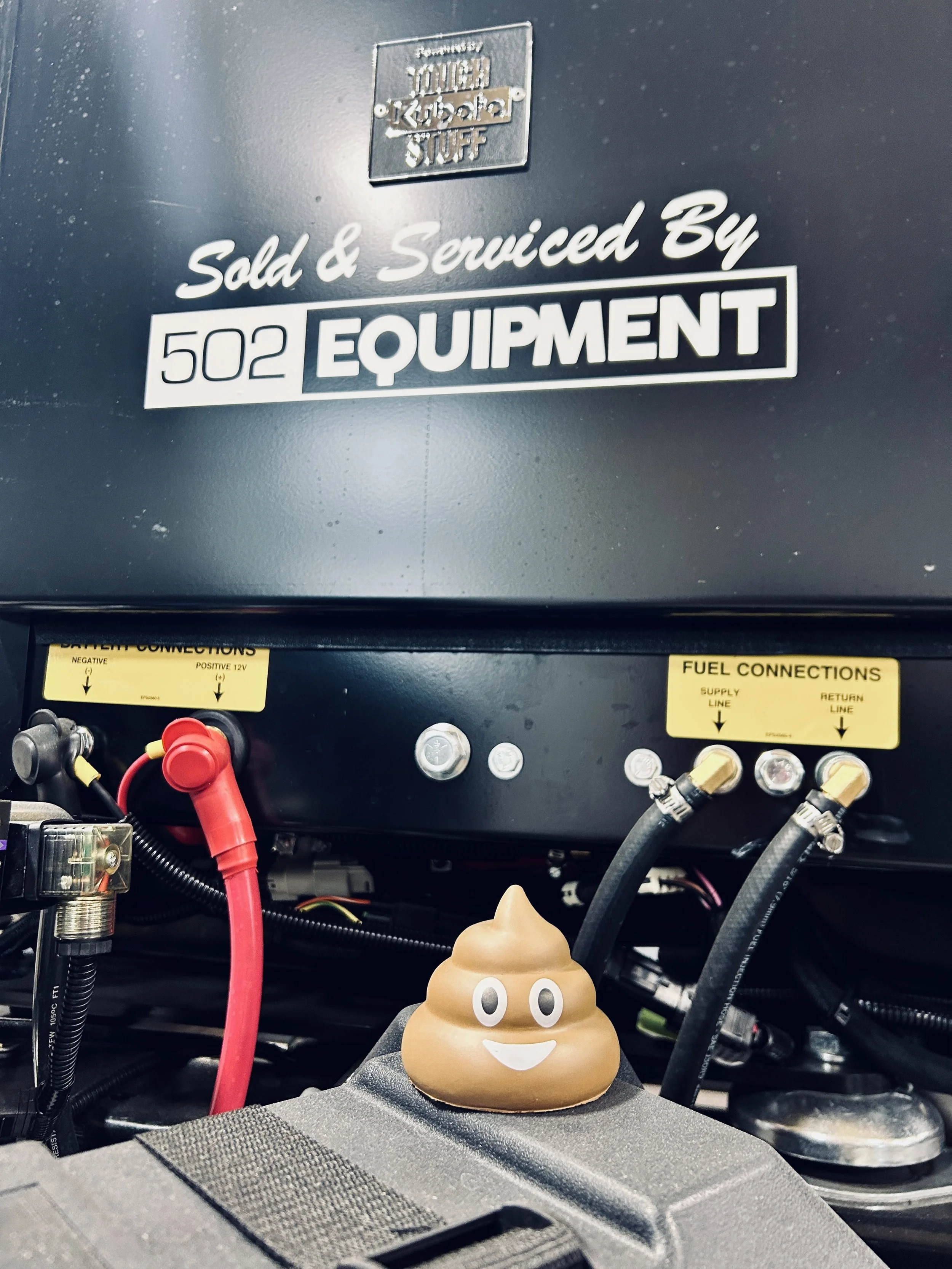 A small poop emoji plush with cartoon eyes and a smiling face placed on a mechanical surface, with a background of electrical wires and equipment labeled 'Sold & Serviced By 502 Equipment' and various electrical connections.