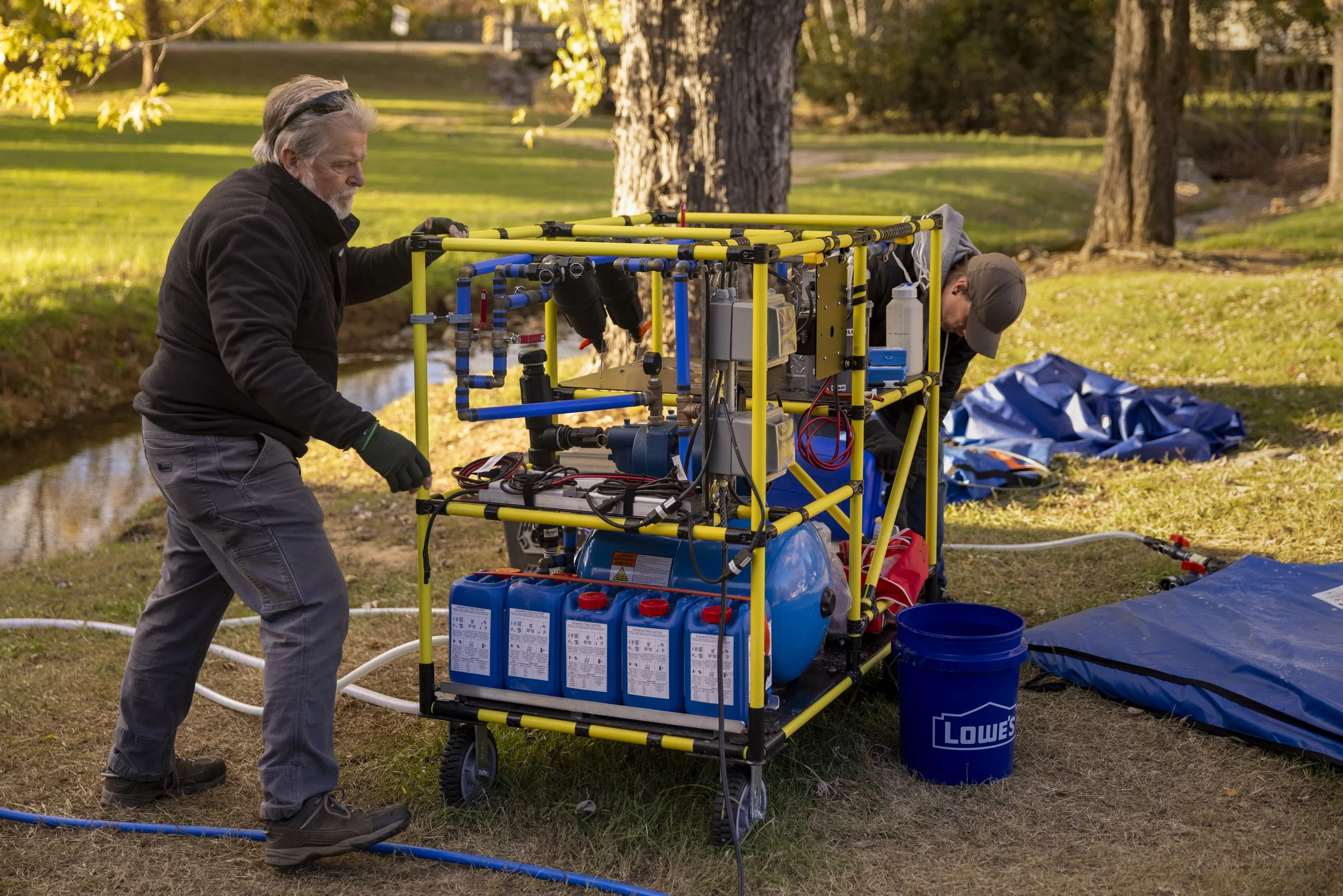 Two men working with the WaterStep WOWCart mobile wate  purification system, outdoors near a Murfreesboro, Kentucky river with trees and grass, with supplies and equipment around them.