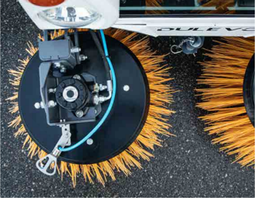 Close-up of a dulevo street sweeper's rotating brush with orange bristles on a paved surface, part of a larger cleaning machine, partly visible.