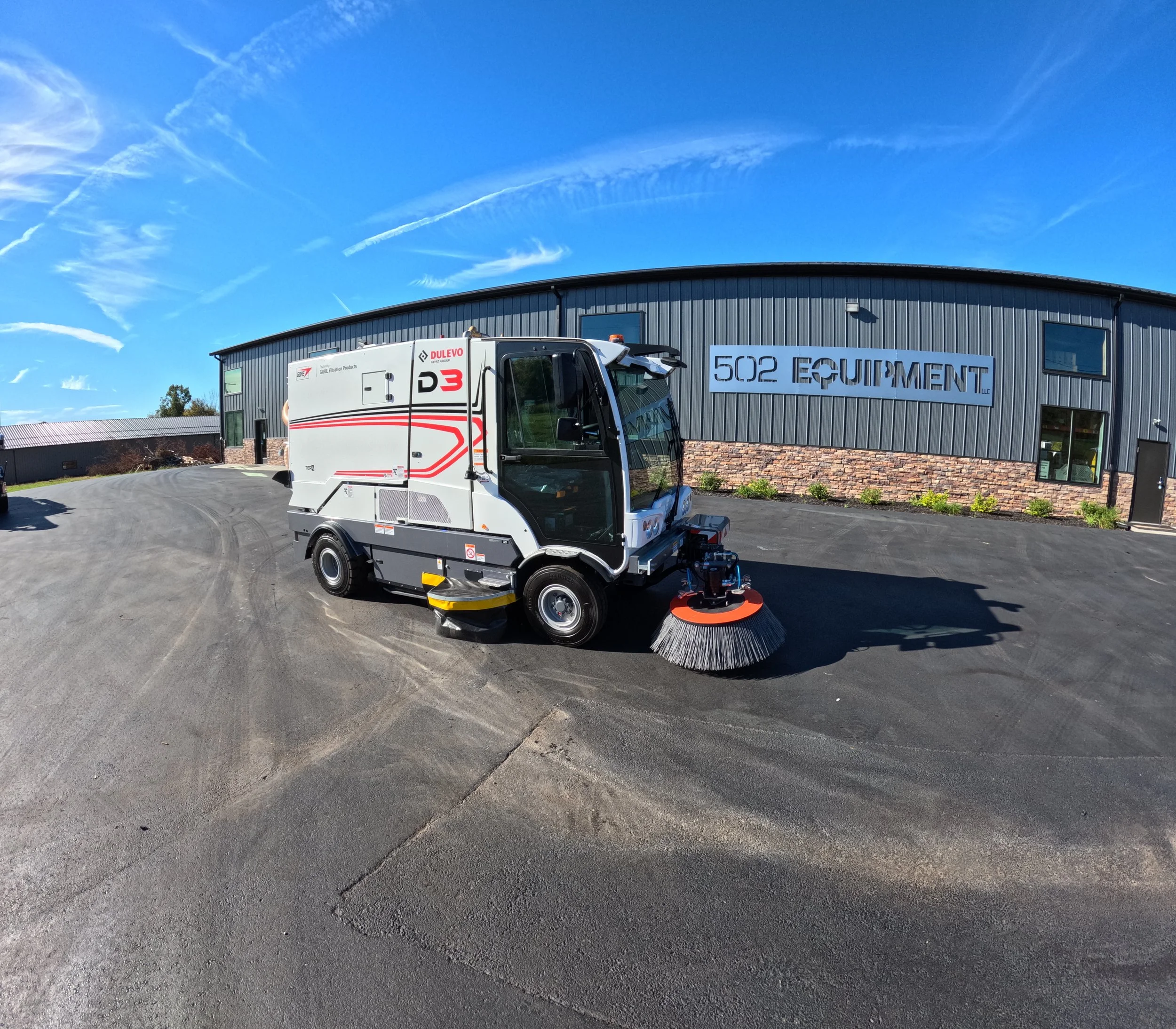 A Dulevo street sweeper vehicle on a paved lot in front of a building with a sign that reads '502 Equipment' under a clear blue sky.