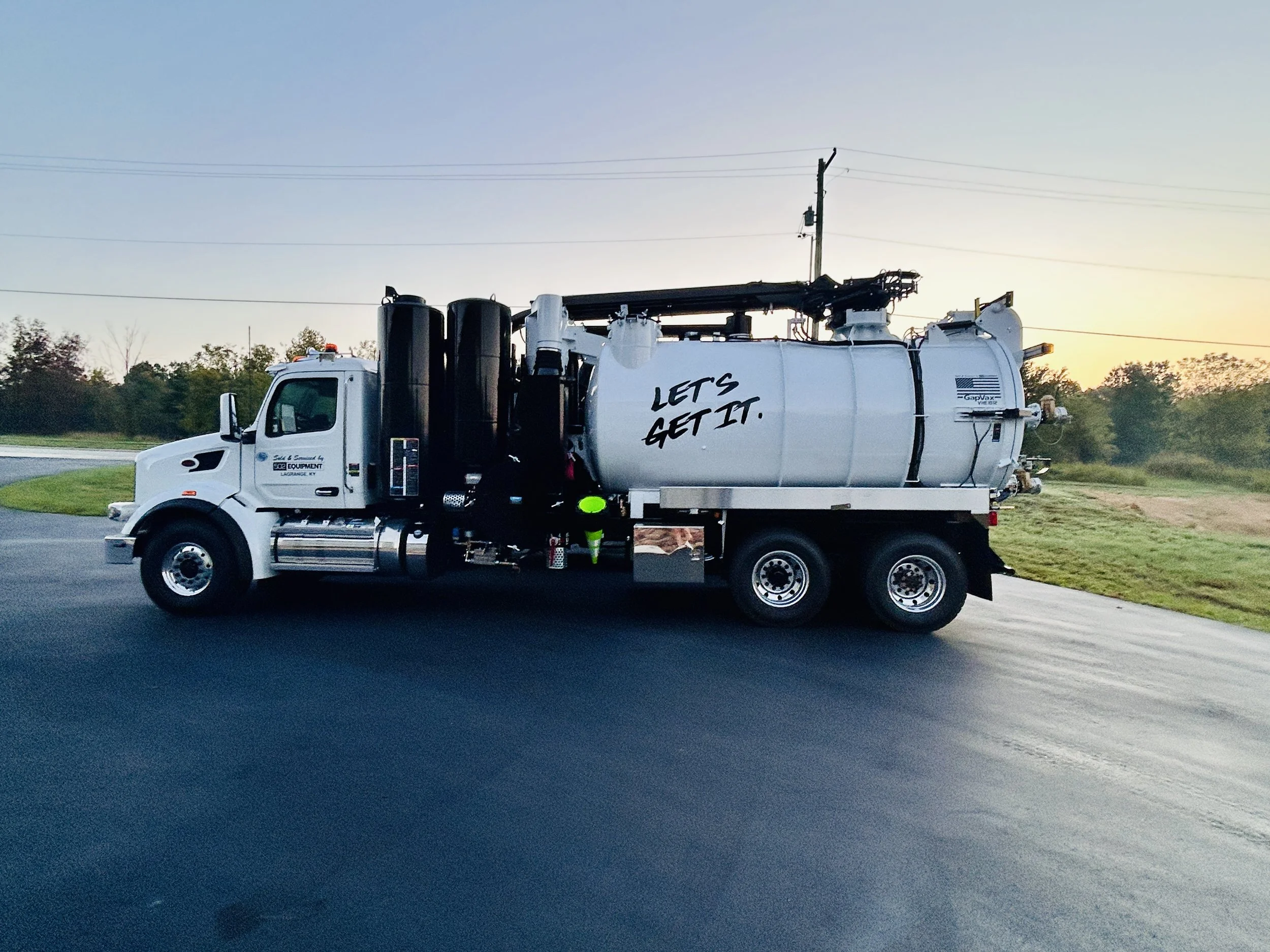 A large white GapVax VHE vacuum truck with the words "Let's Get It" written on the side, parked on the side of a paved road at sunset in Mayfield, Kentucky, with trees and power lines in the background.