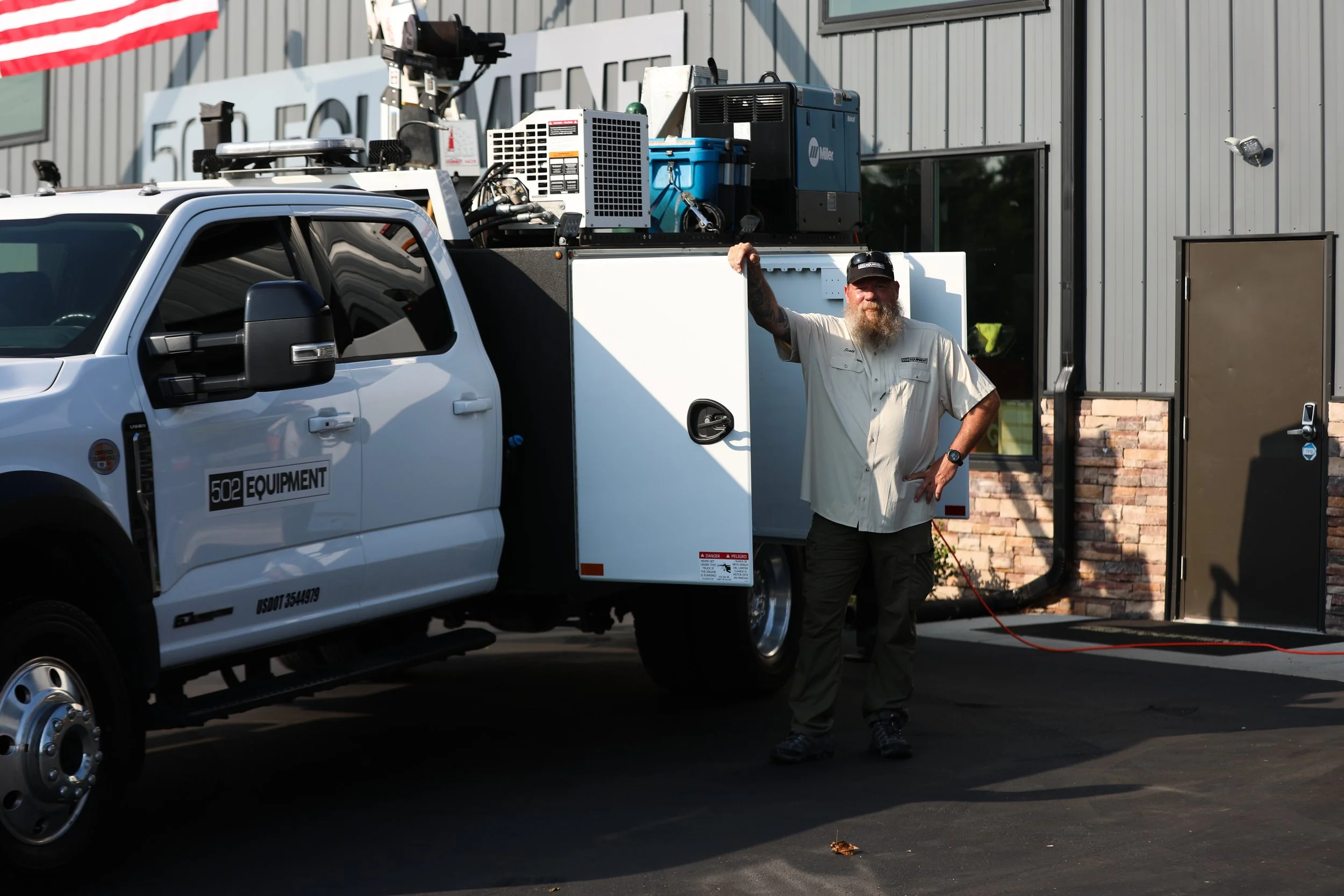 Scott Brinkman, master mechanic is is smiling and leaning against his mobile service truck with one arm raised. The truck is parked outside the 502 Equipment service center in Lagrange, KY