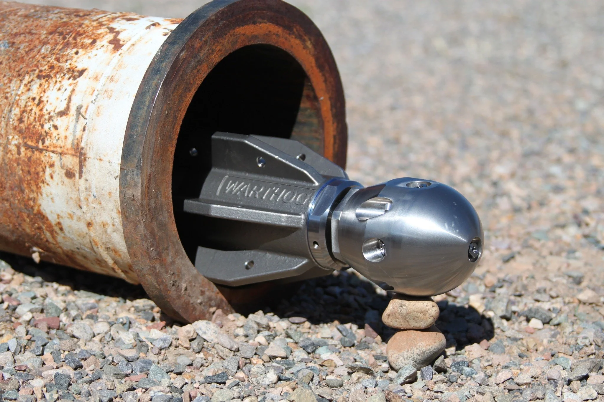 A large, rusty metal pipe on a gravel surface, with a Warthog Sewer Nozzles partially inside it. The nozzle has the word 'WARTHOG' engraved on its side and is supported by small rocks underneath.