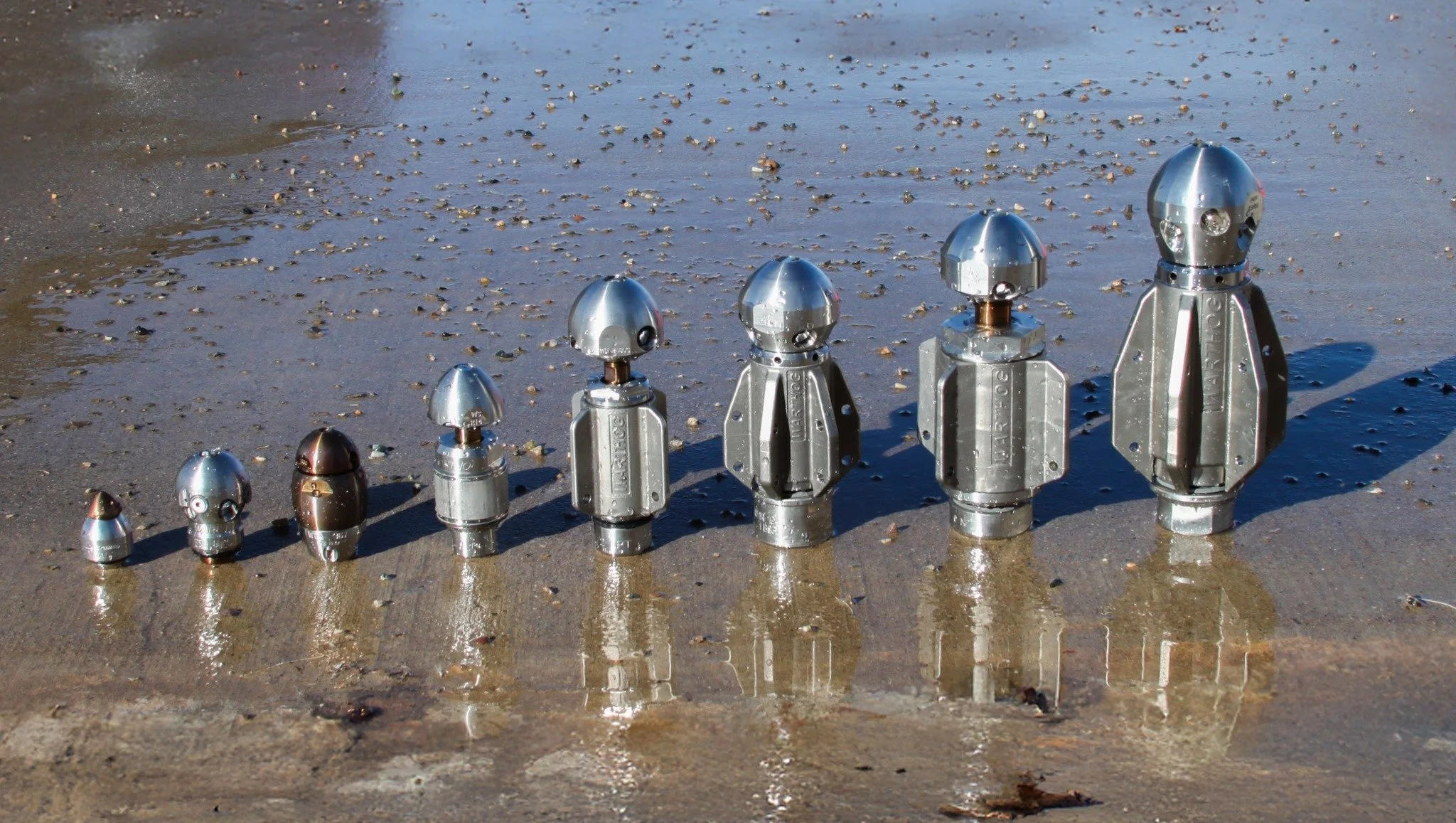 Series of seven metal Warthog sewer jetting nozzles near the water, progressively increasing in size from left to right.