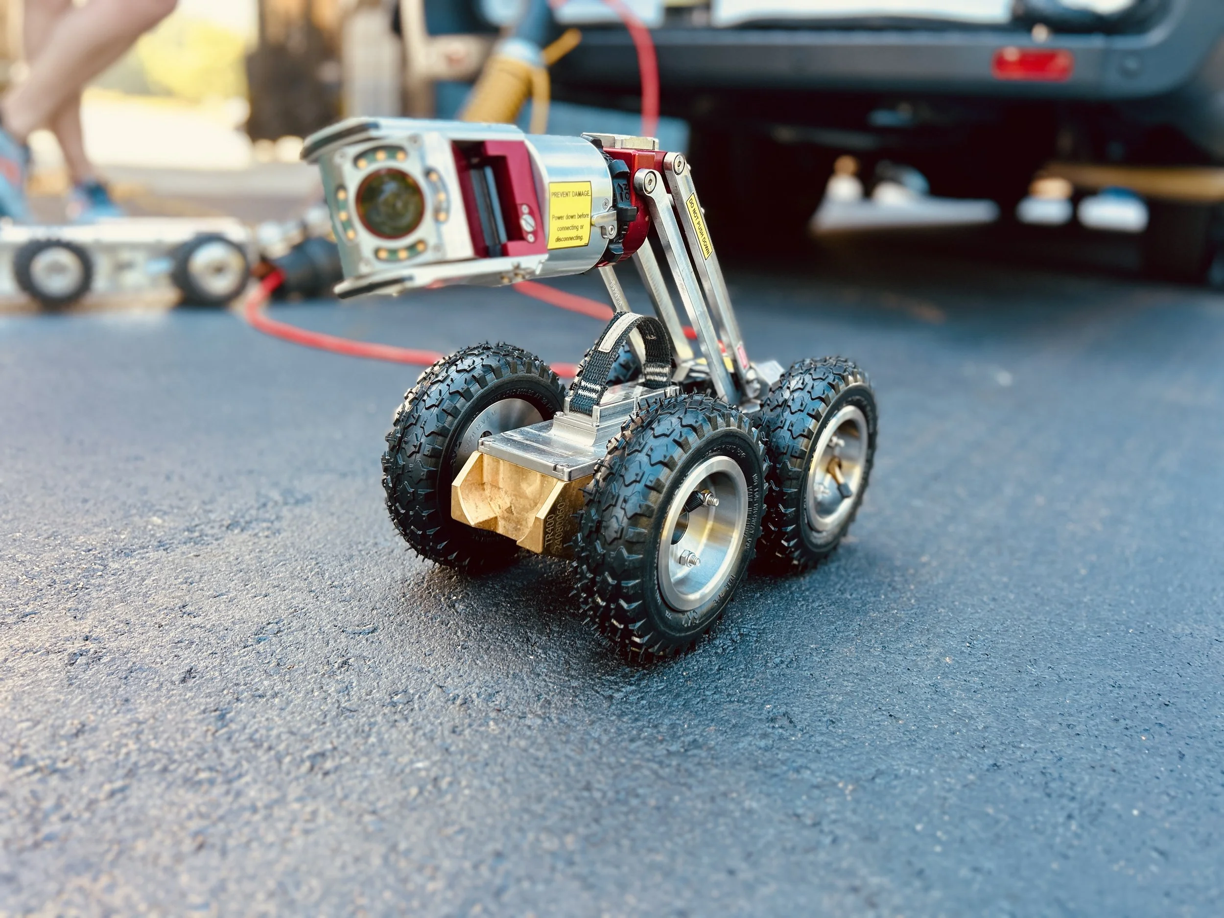 Close-up of a small robotic device equipped with a camera and lights, the Aries Voyager mainline pipe inspection system, positioned on asphalt with a utility truck and houses in the background near Evansville, Indiana.