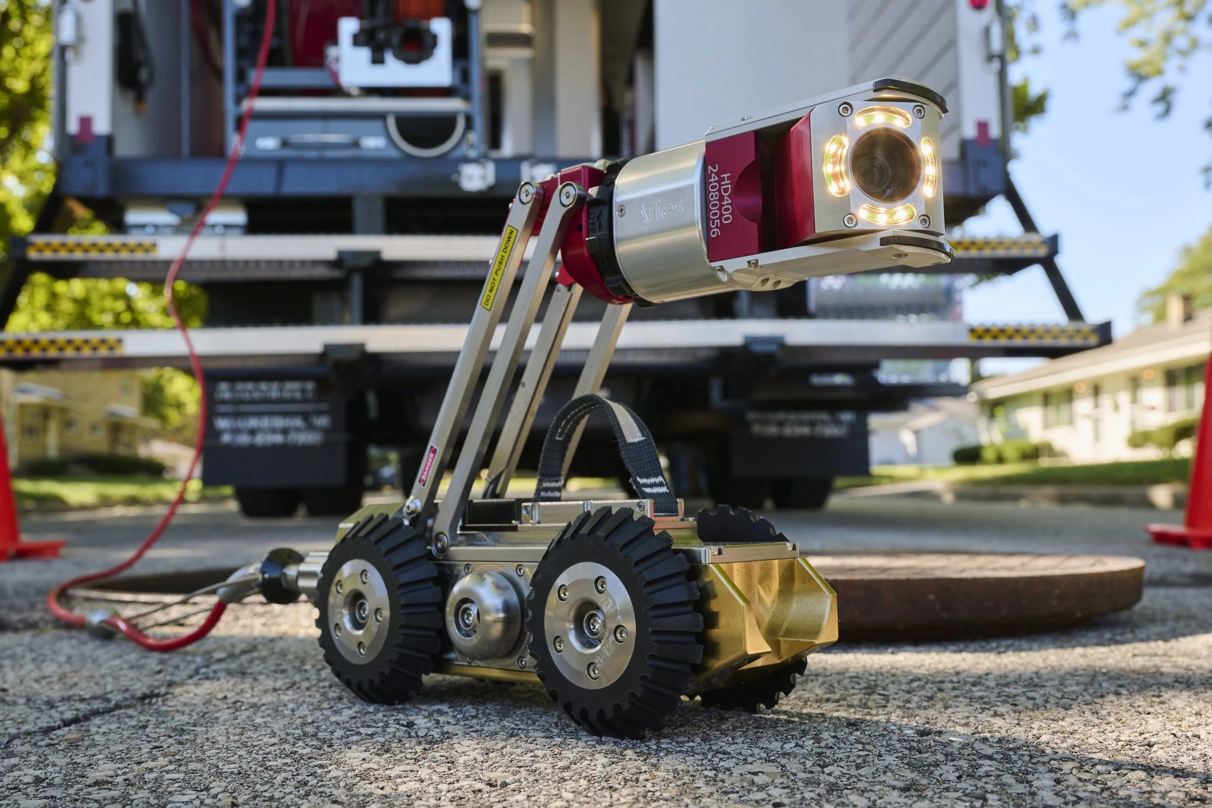 Close-up of a small robotic device equipped with a camera and lights, the Aries Voyager mainline pipe inspection system, positioned on asphalt with a utility truck and houses in the background near Evansville, Indiana.