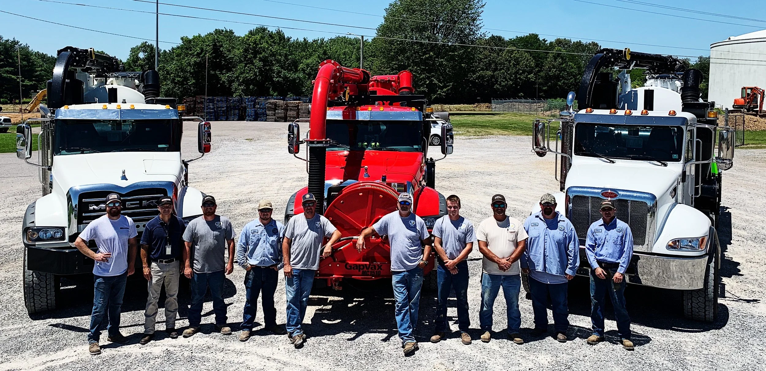 Ten men standing in front of three large vacuum trucks trucks, two white and one red, on a gravel lot with trees and a blue sky in the background in Mayfield, KY