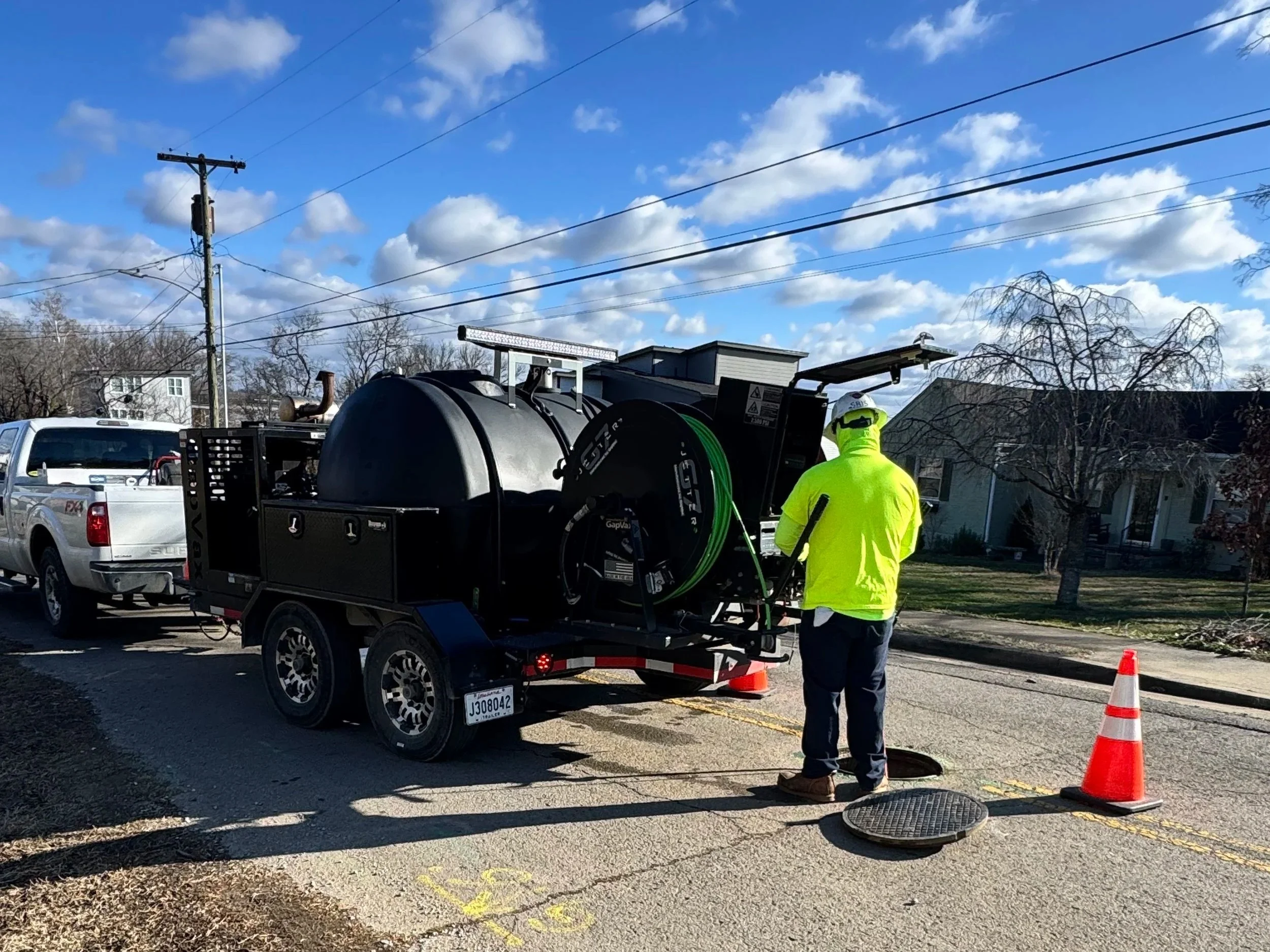 Worker in yellow safety vest jetting pipes with a GapVax g7 trailer jetter on suburban street with orange traffic cones and a manhole cover open.