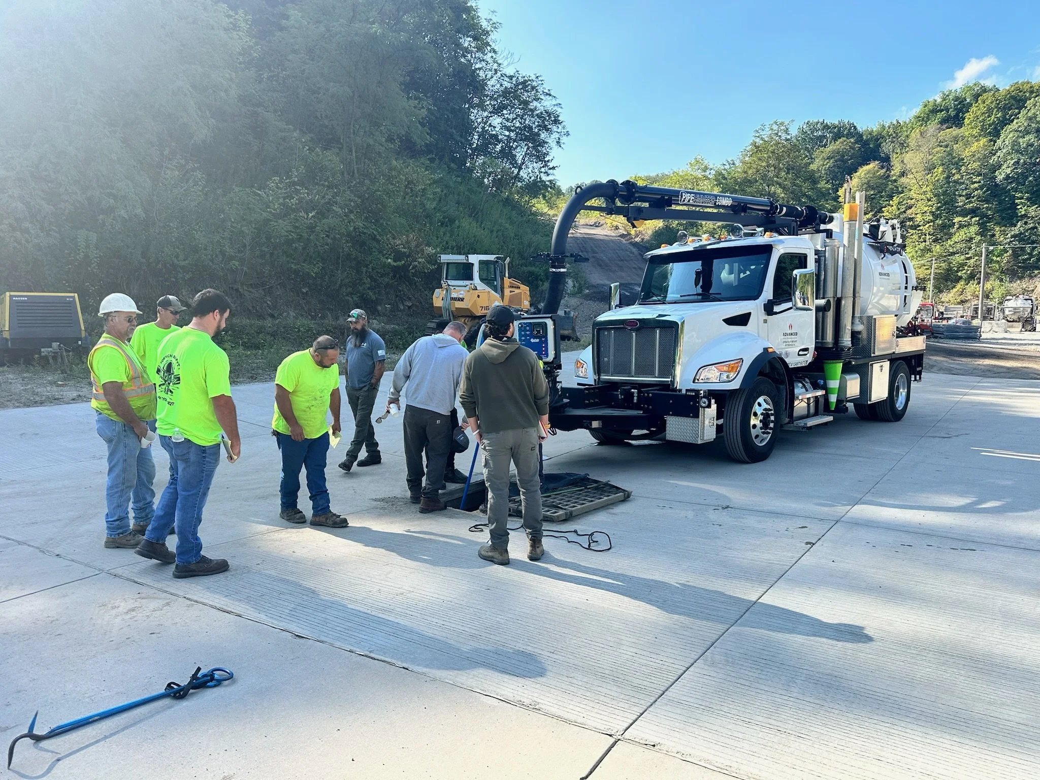 Group of construction workers and engineers gathered around a manhole at a construction site. There is a large GapVax vacuum truck nearby.