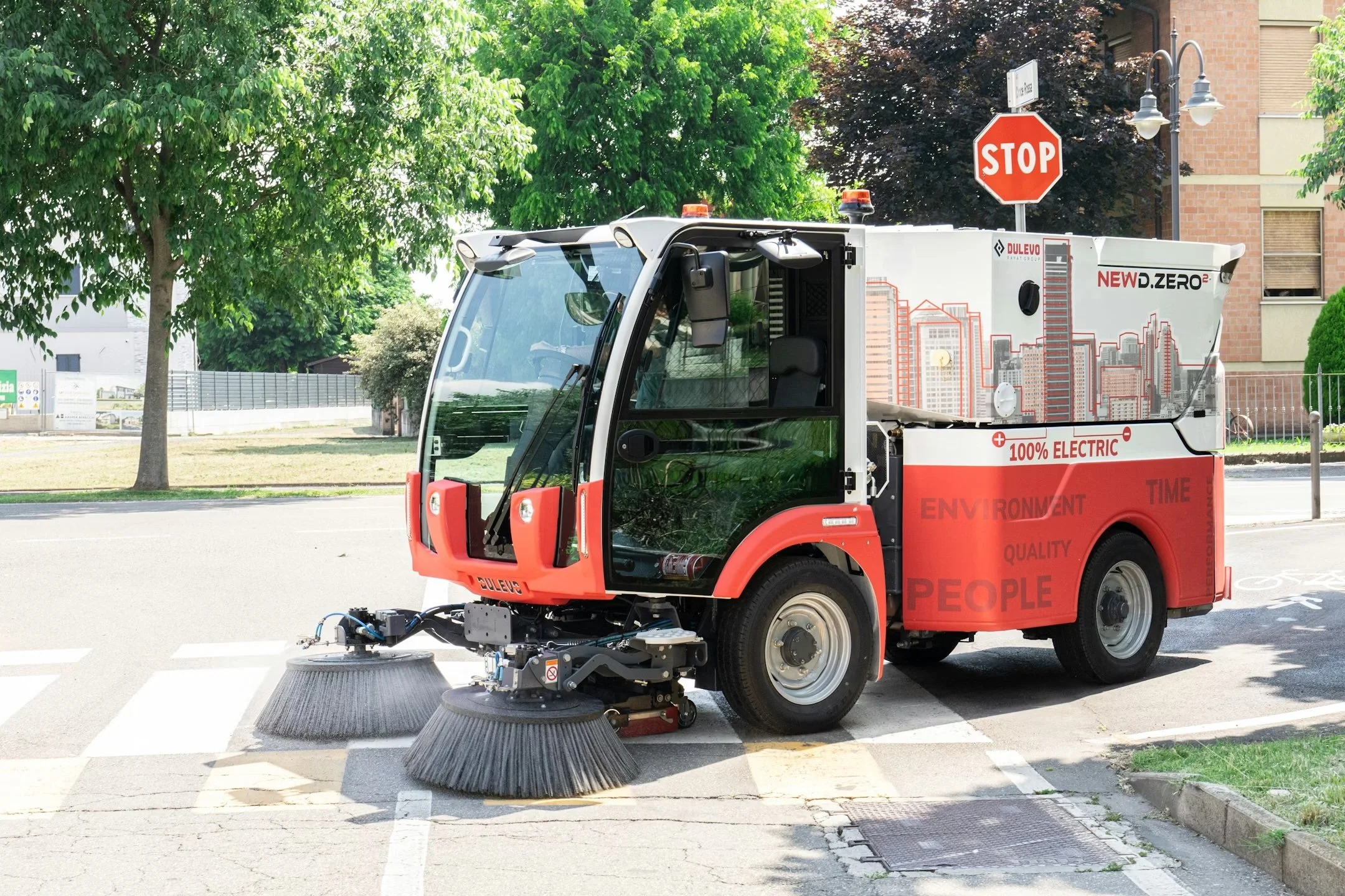 A Dulevo DZero all electric street sweeper sold by 502 Equipment in Kentucky, Indiana and Tenessee parked on a city street, with large brushes at the front and a cityscape illustration on its side.