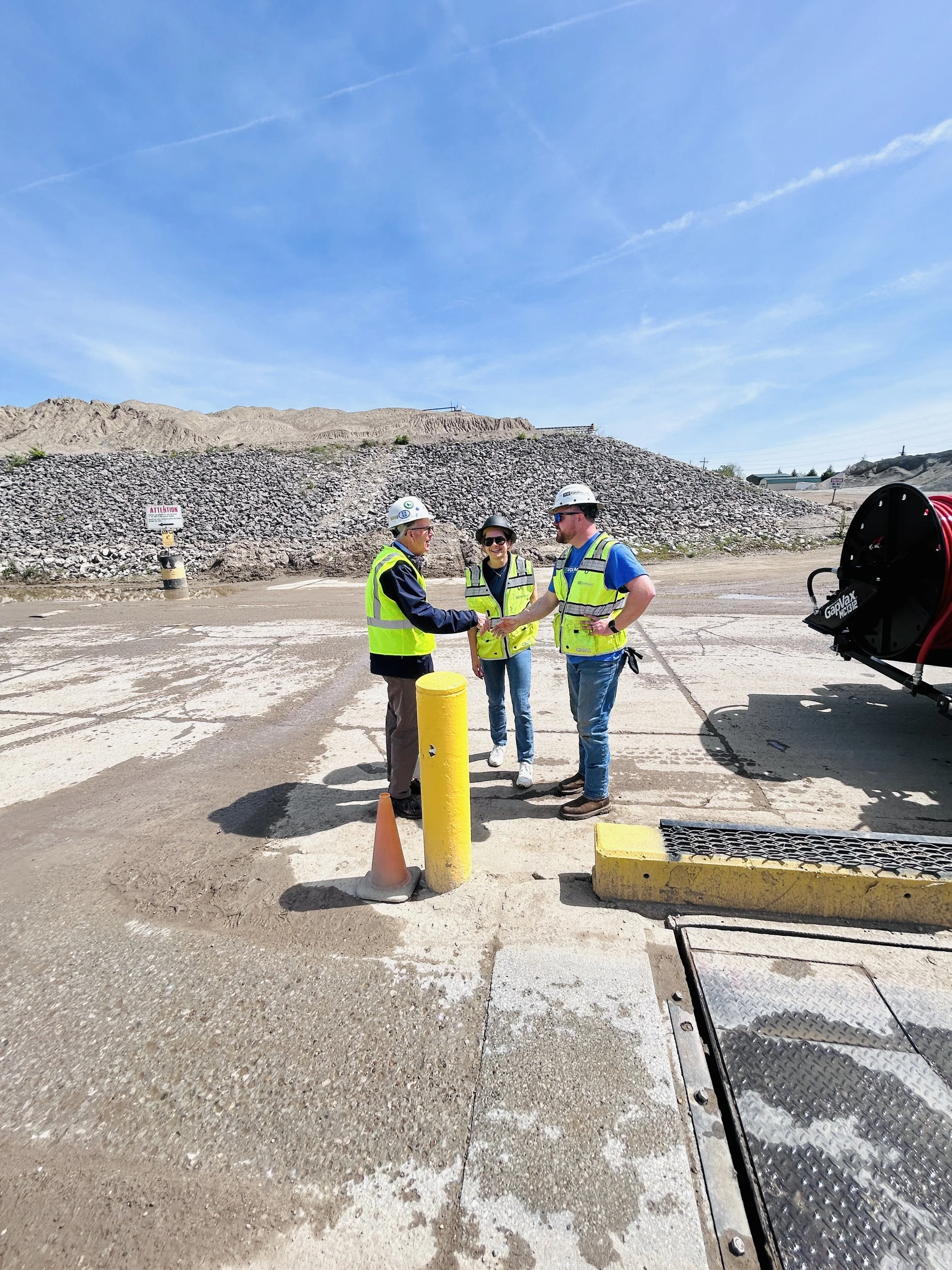 Three construction workers shake hands outdoors at a construction site, wearing safety vests and helmets with a dirt and gravel landscape in the background.