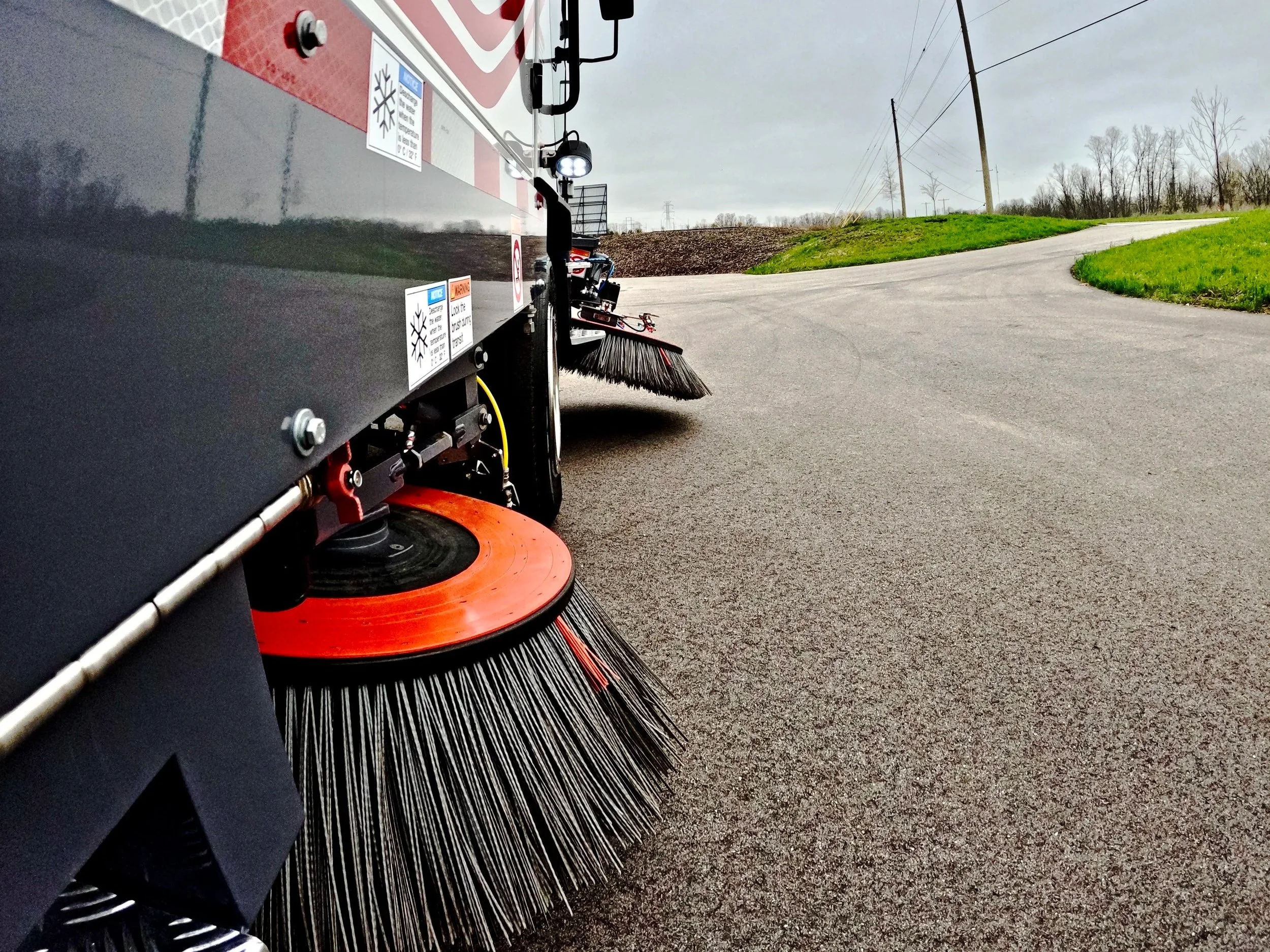 Close-up view of dulevo street sweeper brushes sweeping a paved road with grassy area and power lines on a cloudy day near Lexington, Kentucky.