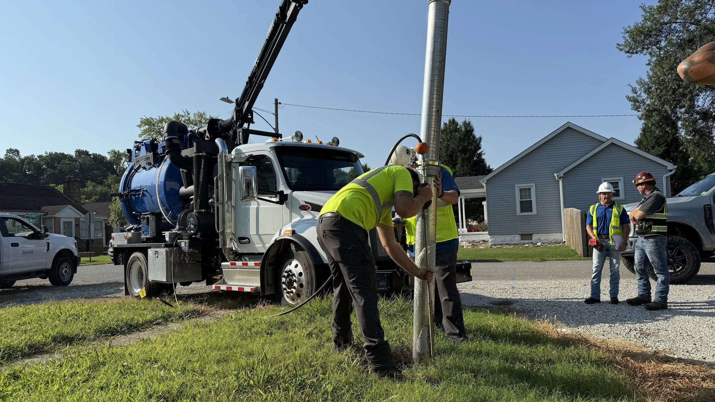 Workers in yellow and blue vests and hard hats using a vacuum truck to daylight utilities in a residential area. 
