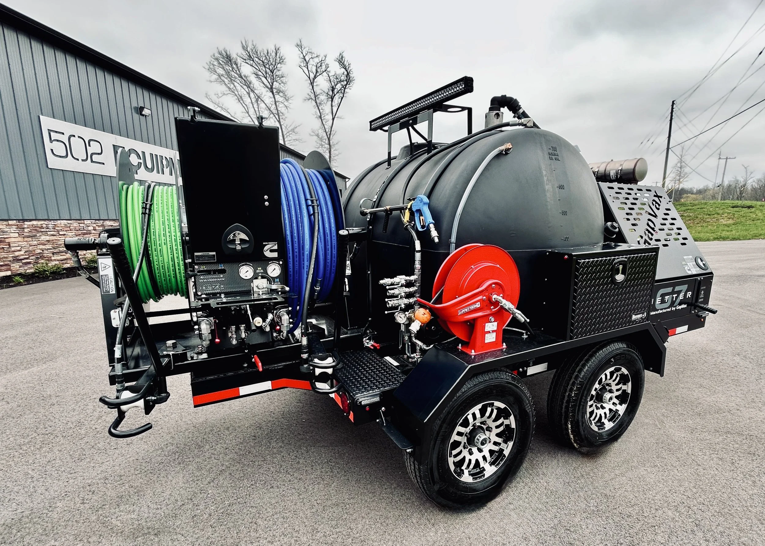 A GapVax G7 dual reel trailer jetter with two wheels, featuring hoses, a pressure gauge, and a large black tank, parked on an asphalt surface near LaGrange, KY and trees in the background.