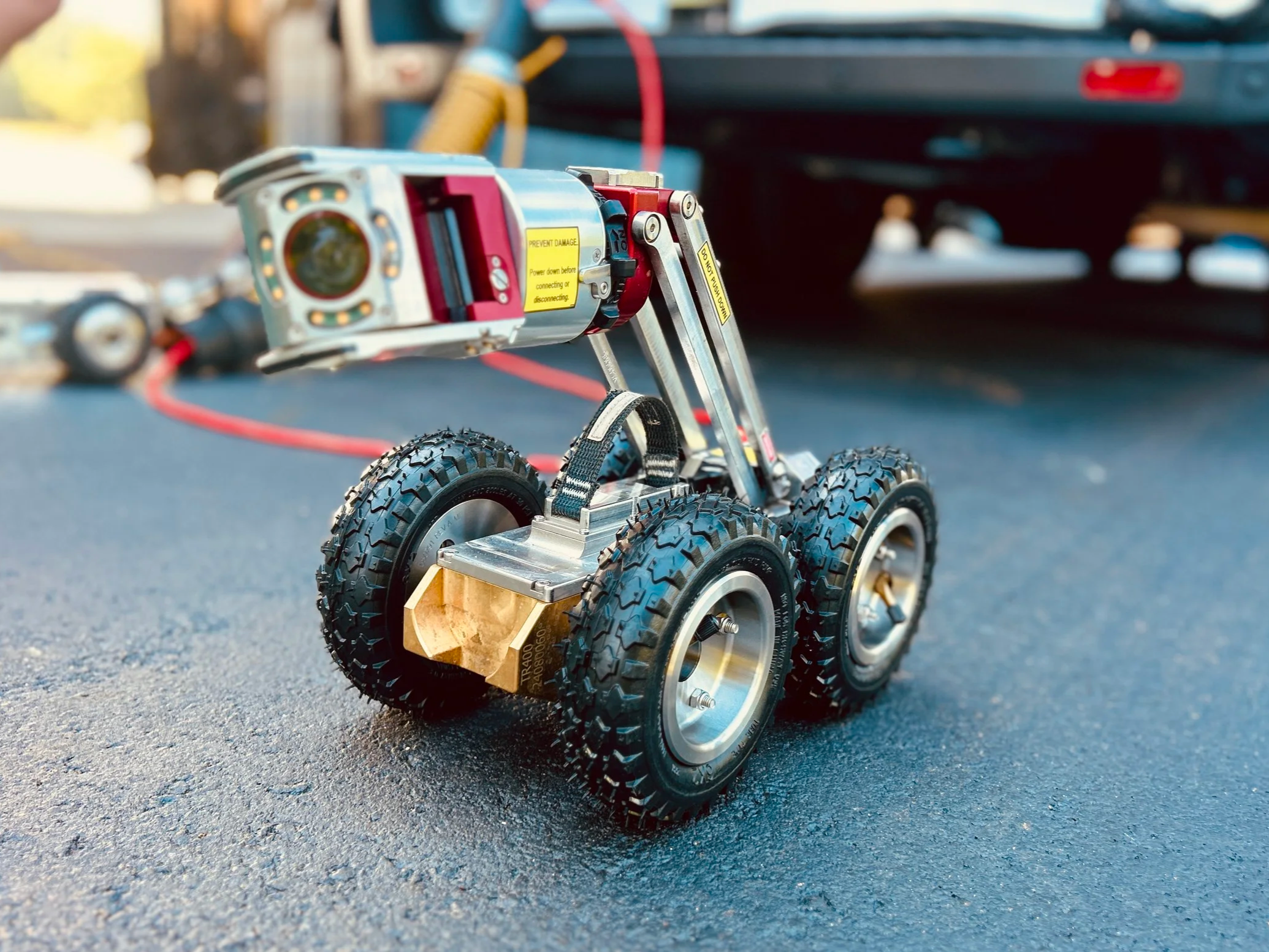 Close-up of a small robotic device equipped with a camera and lights, the Aries Voyager mainline pipe inspection system, positioned on asphalt with a utility truck and houses in the background near Evansville, Indiana.