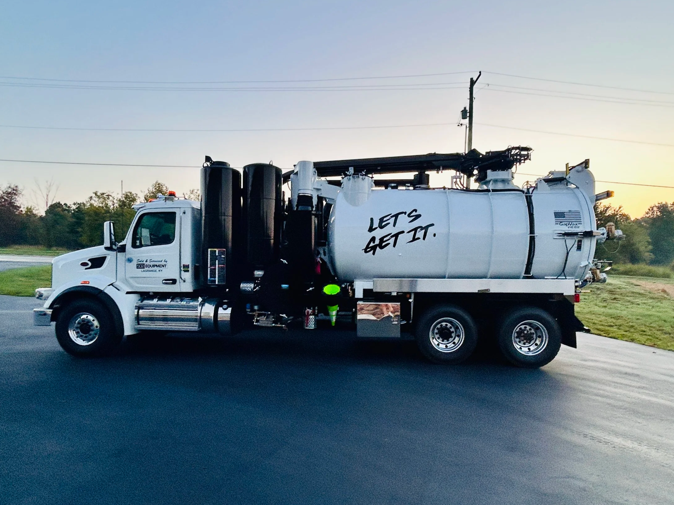 A white GapVax VHE vacuum truck, sold by 502 Equipment with the slogan "LET'S GET IT" written on the tank, parked on a paved road with greenery and trees in the background during sunset.