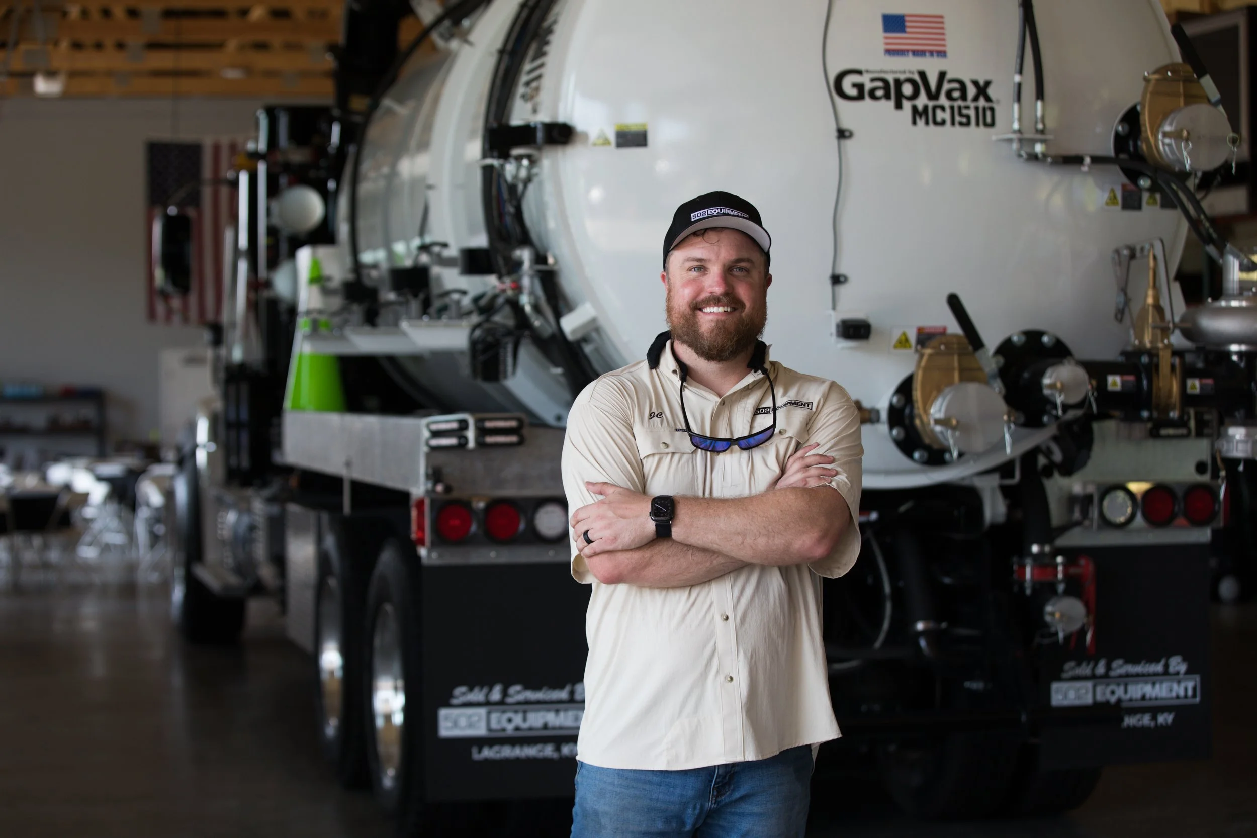 JC Spalding wearing a beige shirt with a logo, sunglasses hanging from his collar, a watch, and a black cap, standing with arms crossed in front of a large white industrial machine labeled "GapVax" inside the 502 equipment service center. 