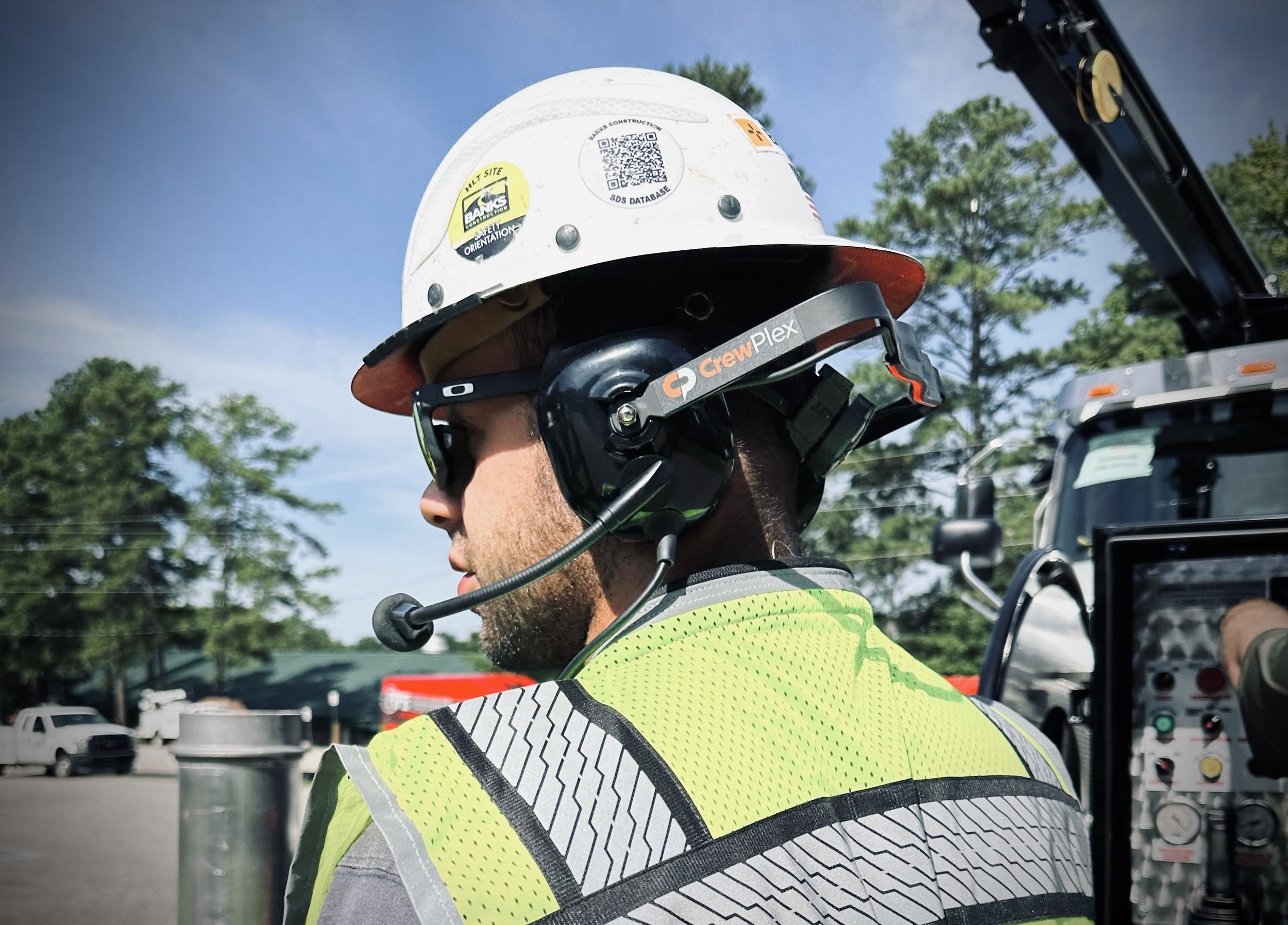 Construction worker wearing a white hard hat, black sunglasses, a Crewplex wireless headset sold by 502 Equipment with microphone, and a high-visibility safety vest, working outdoors with equipment and trees in the background in Louisville, KY